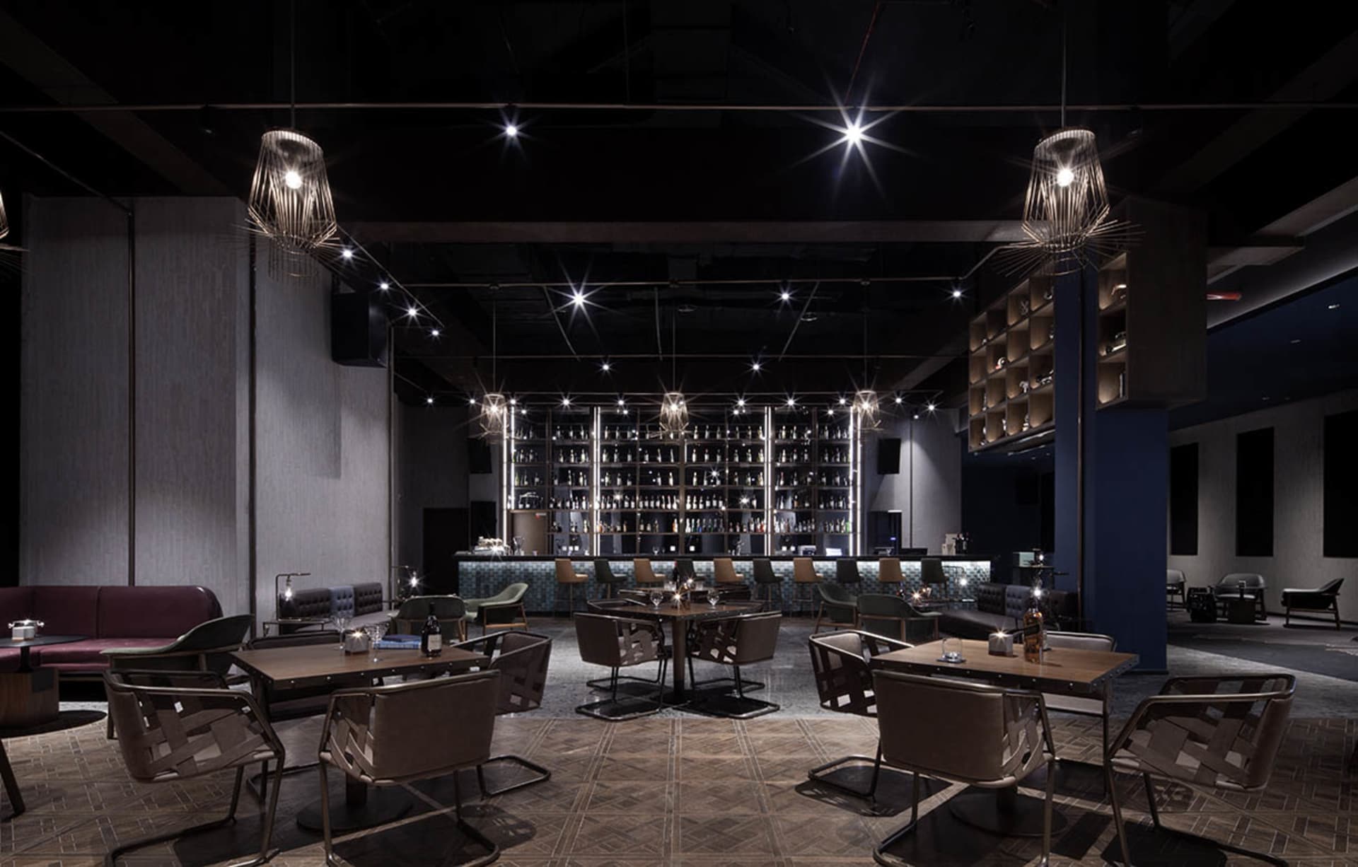 A modern restaurant bar with concrete walls and leather seating, featuring a well-lit central area with tables and chairs.