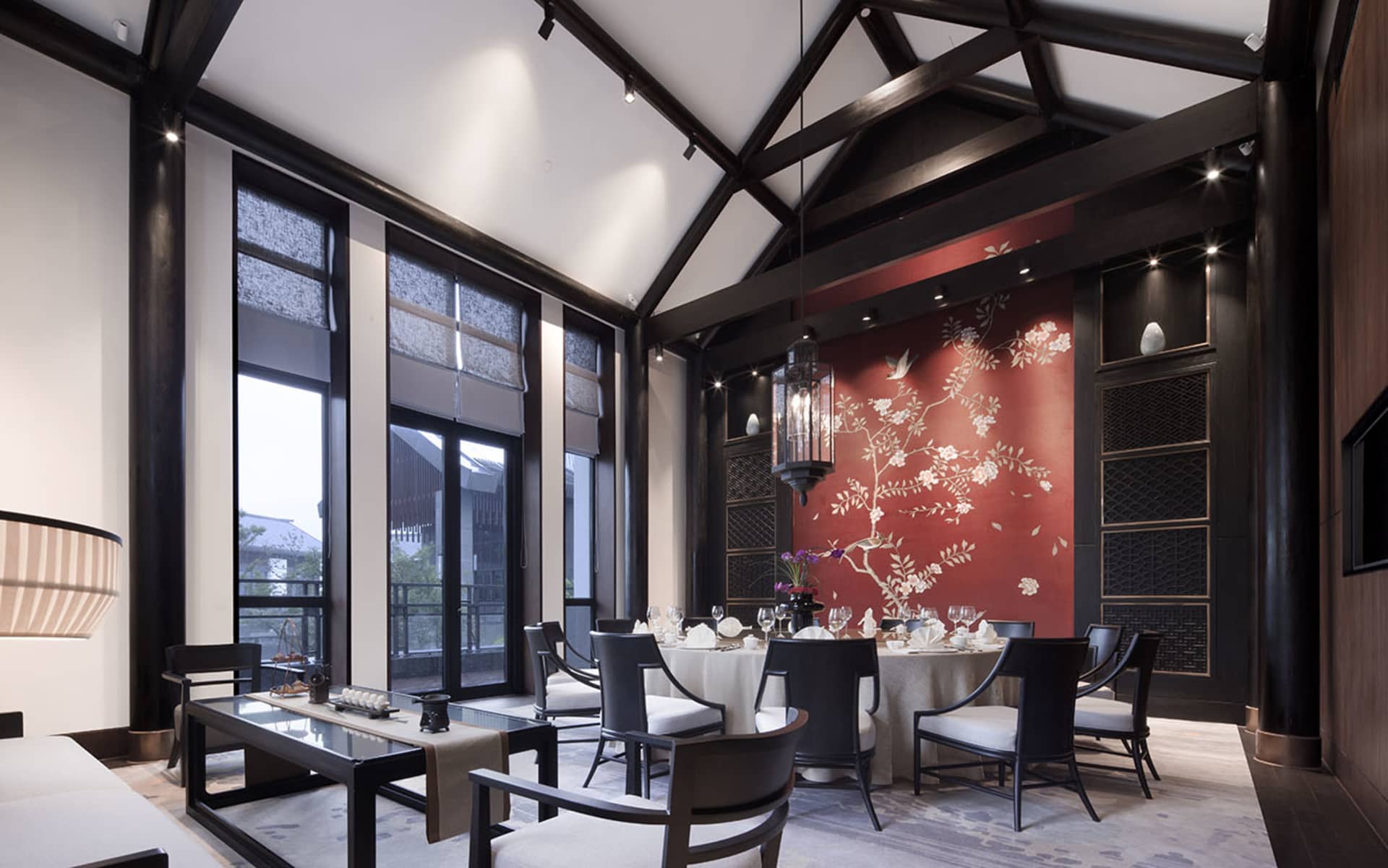 A modern restaurant dining room with a dark wooden ceiling and walls, natural light from large windows, and a round dining ta