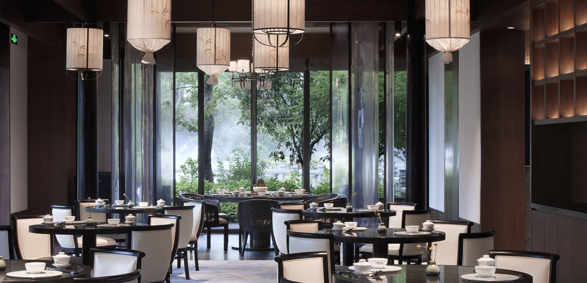 A modern restaurant interior with elegant lighting fixtures and large windows showcasing greenery.