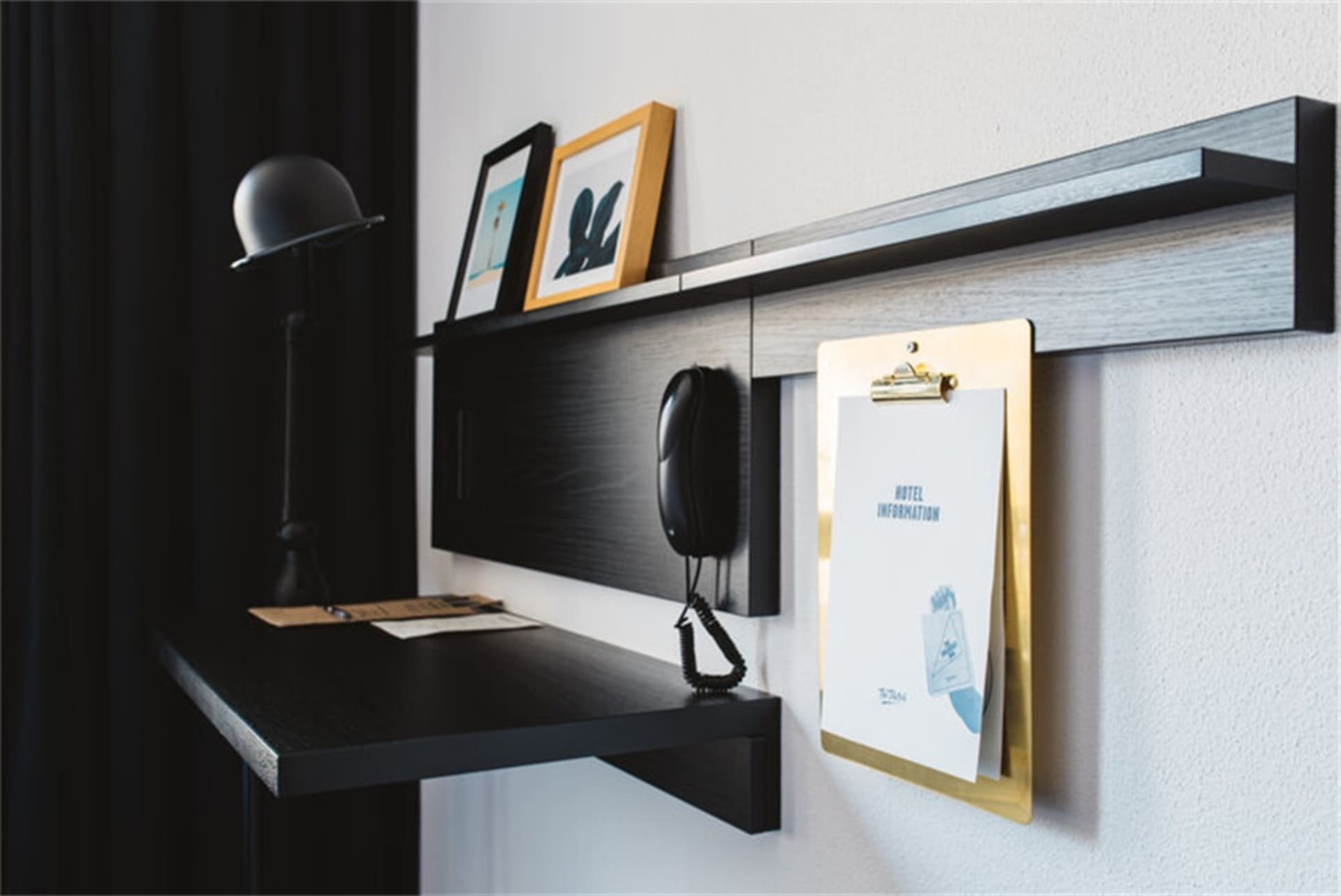 A minimalist hotel room desk with a black desk, black lamp, and a hanging hotel information clipboard.