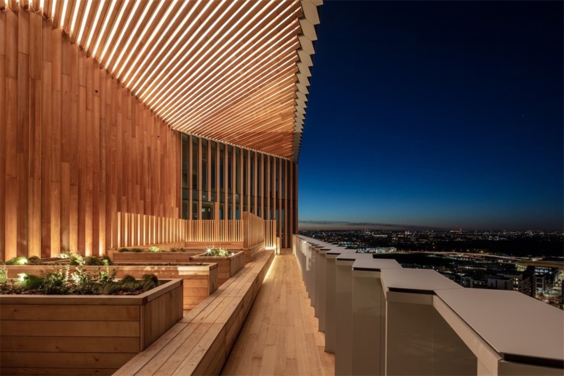 A wooden terrace with planters and a wooden structure at dusk overlooking a cityscape.