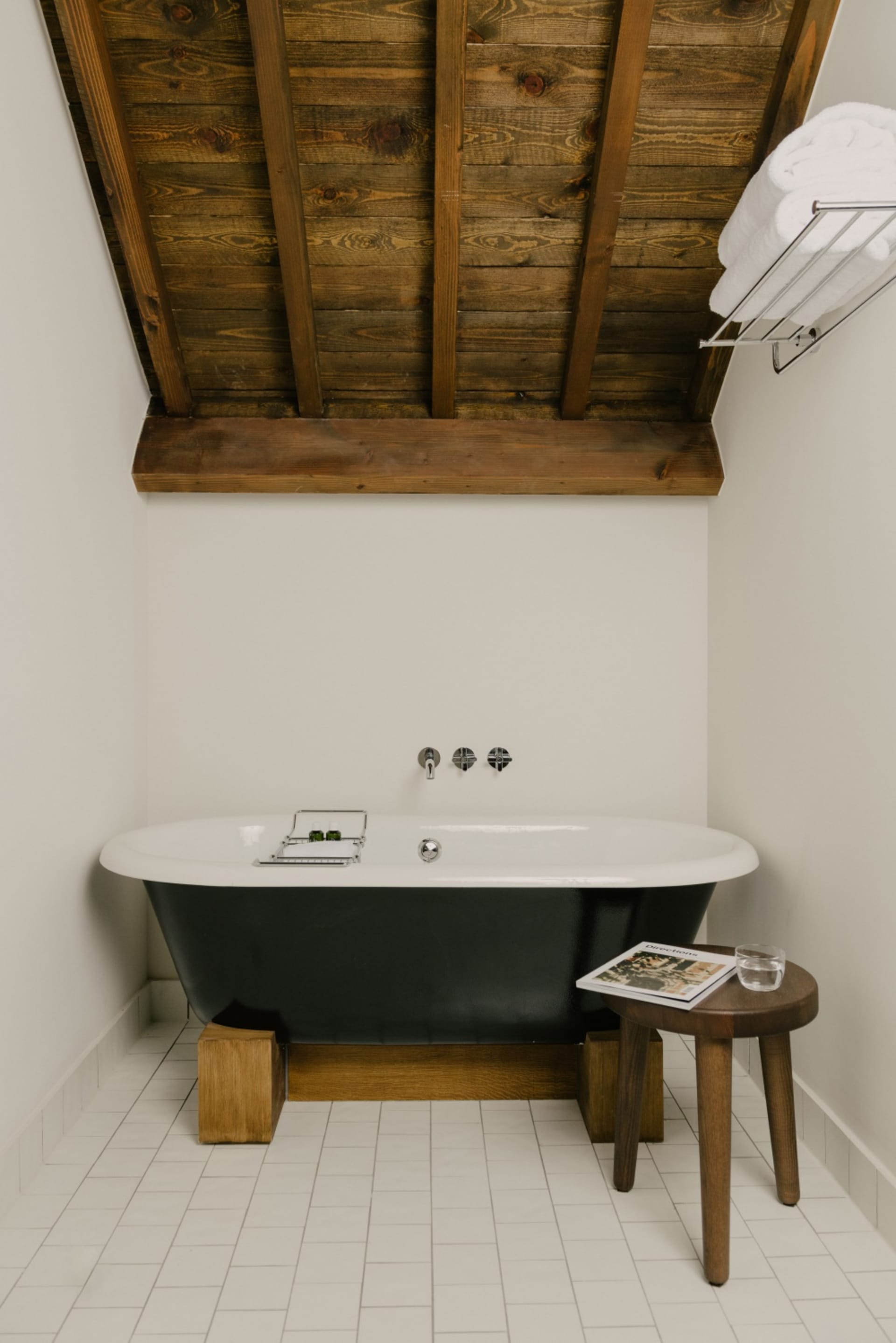 A minimalist bathroom featuring a wooden ceiling with beams, a white tub with black trim, and a wooden stool with a magazine