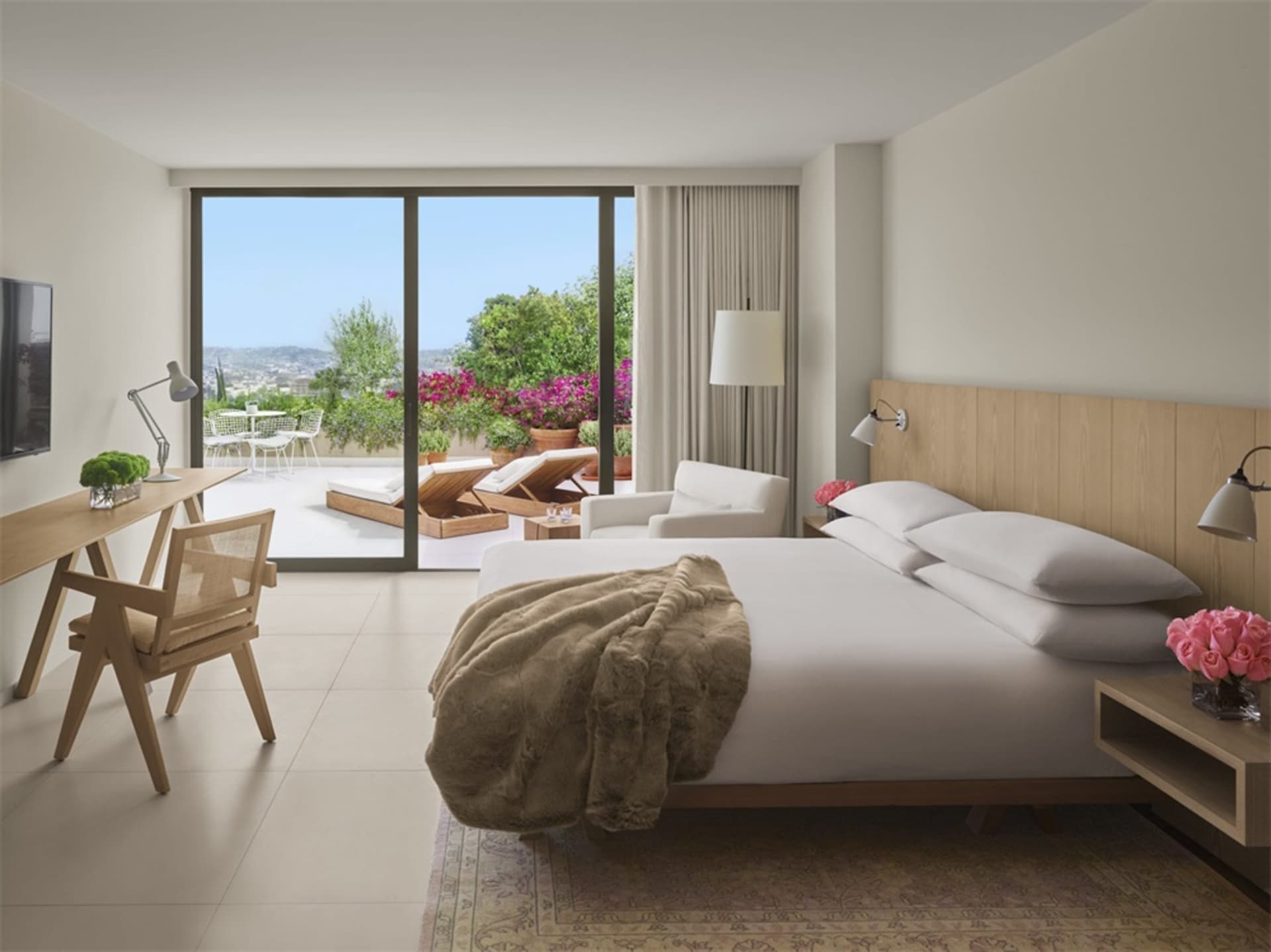 Modern bedroom with white walls, glass door to balcony, wooden bed frame, white bedding, and beige blanket.