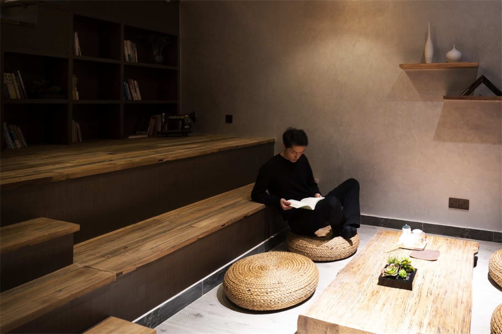 一位人士坐在編織坐墊上，置身於燈光昏黃、木質元素充滿的室內空間，正在閱讀一本書。