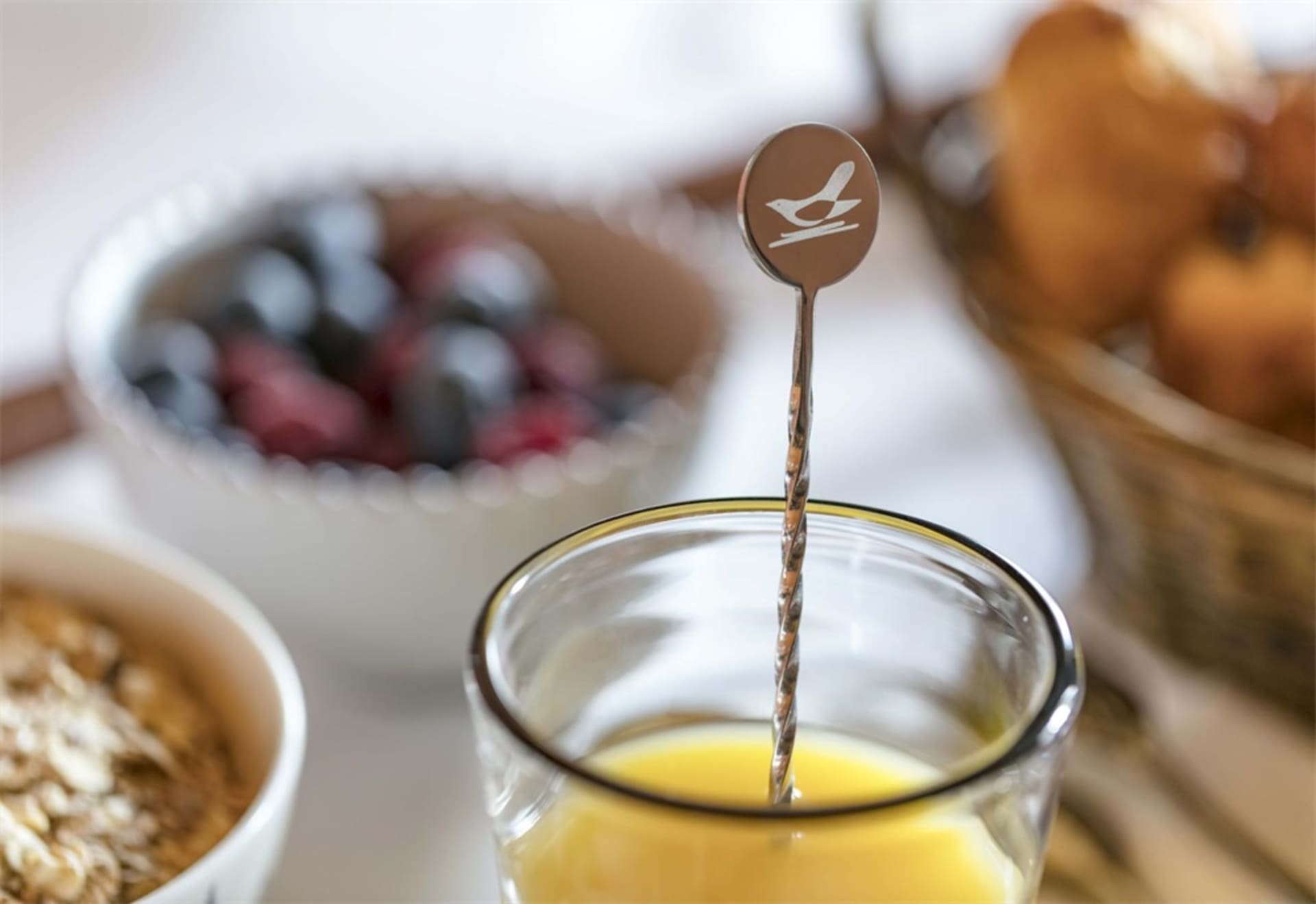 Close-up of a minimalist breakfast scene with a metal stirring spoon in a glass of orange juice, a bowl of mixed berries, and