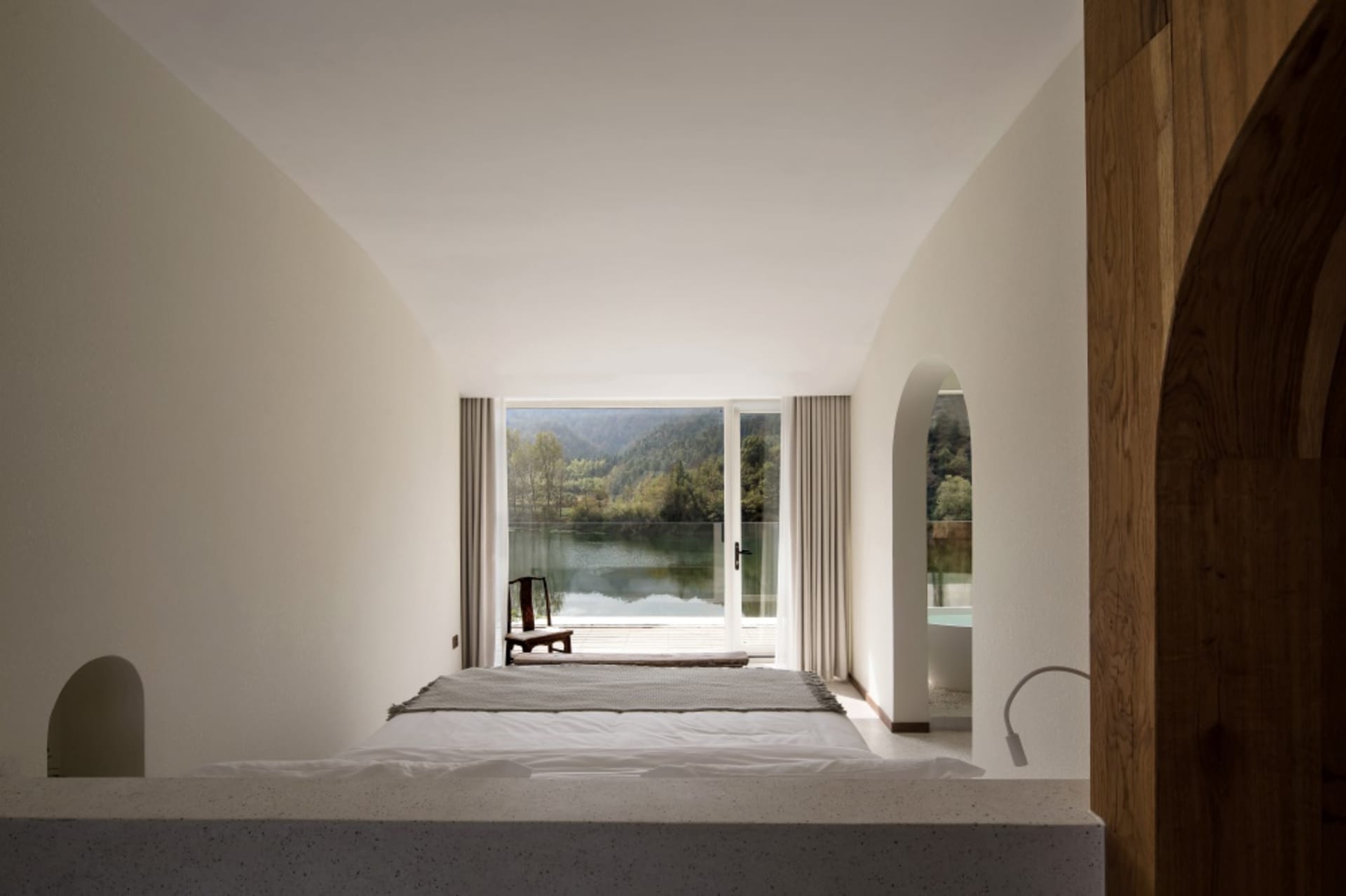 Minimalist bedroom with concrete walls and floor, framed by large windows overlooking a scenic lake view.