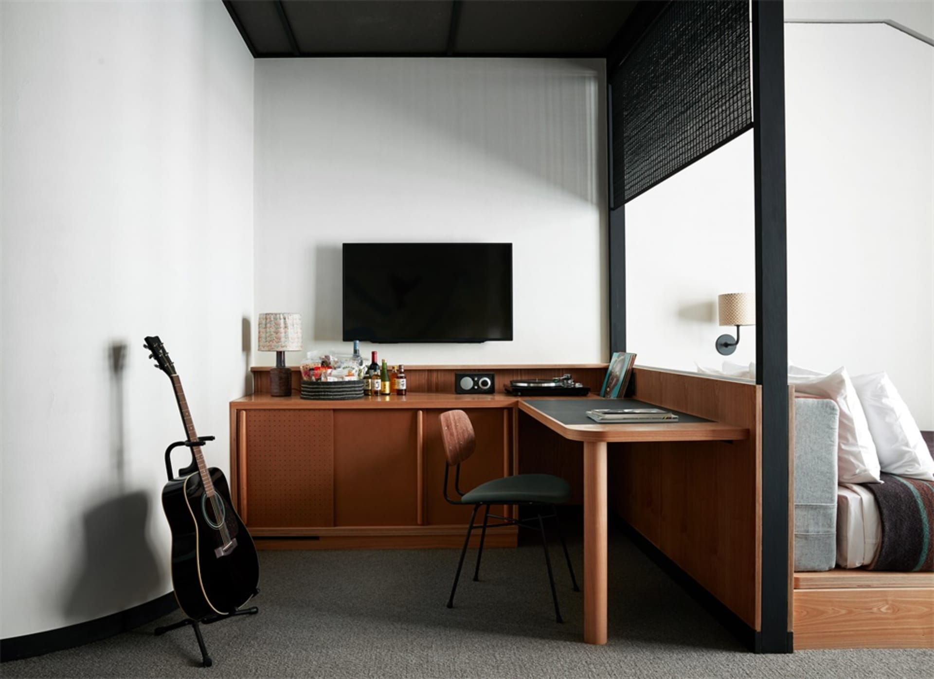 A modern studio apartment featuring a minimalist workspace, a guitar leaning against the wall, and a cozy sleeping area with