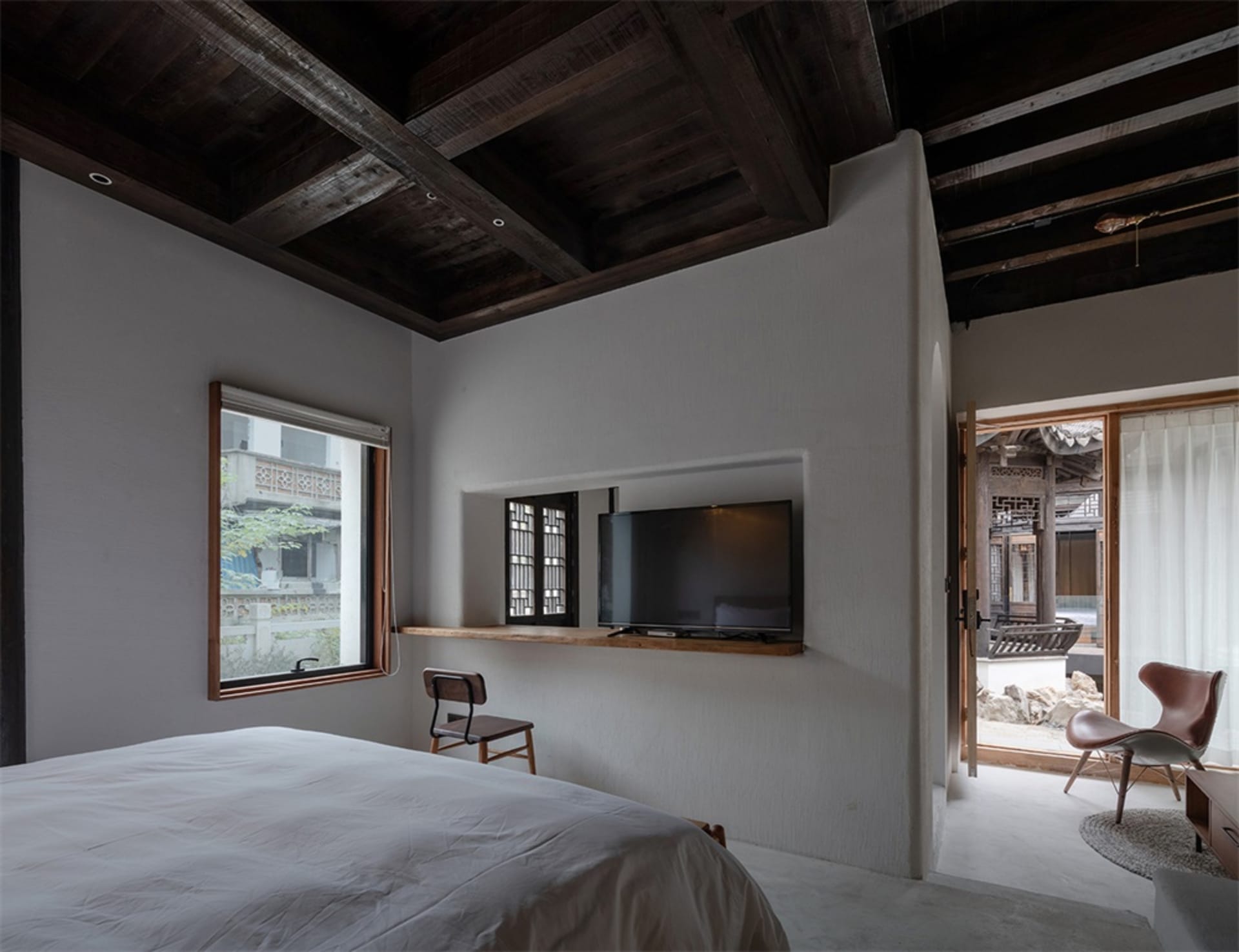 Minimalist bedroom with concrete walls, wood ceiling beams, and a television mounted on a floating shelf.