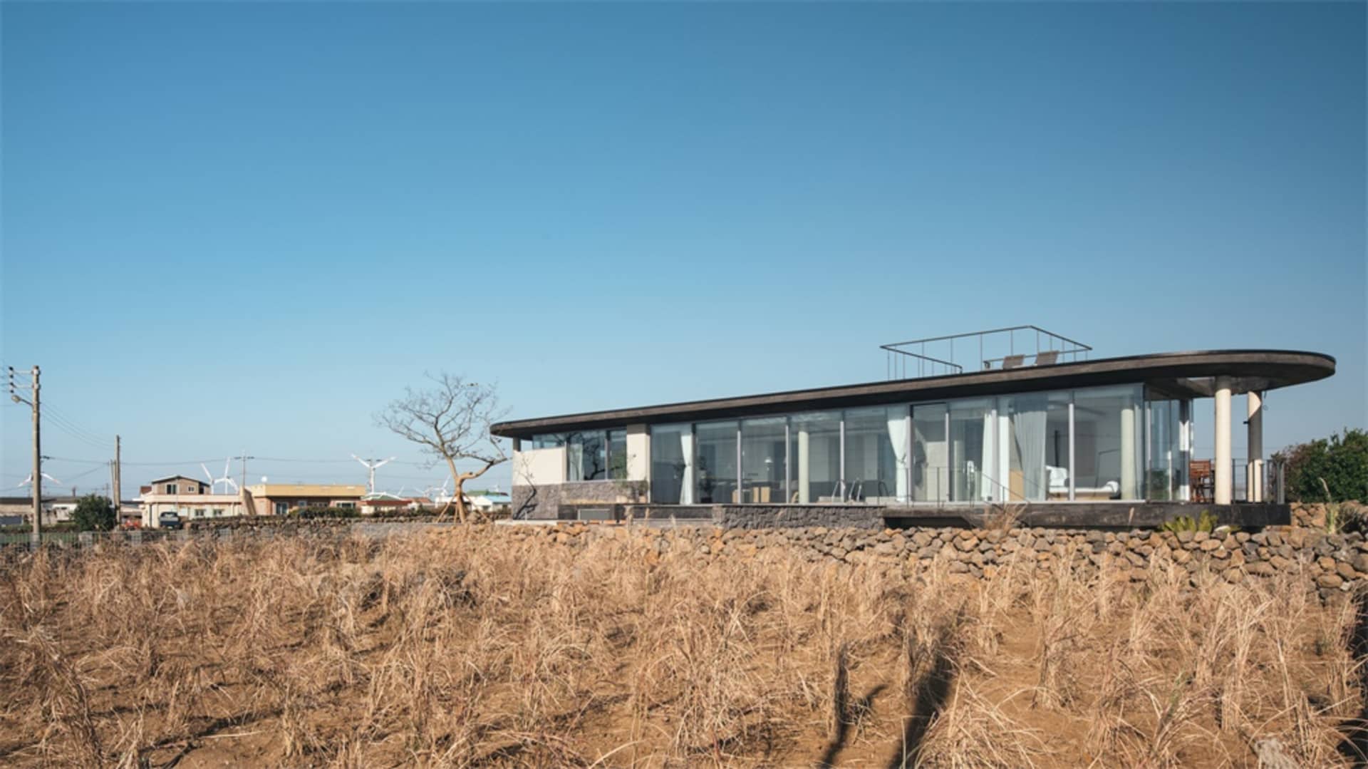 Modern house with glass exterior on a field of dry grass under a clear blue sky.