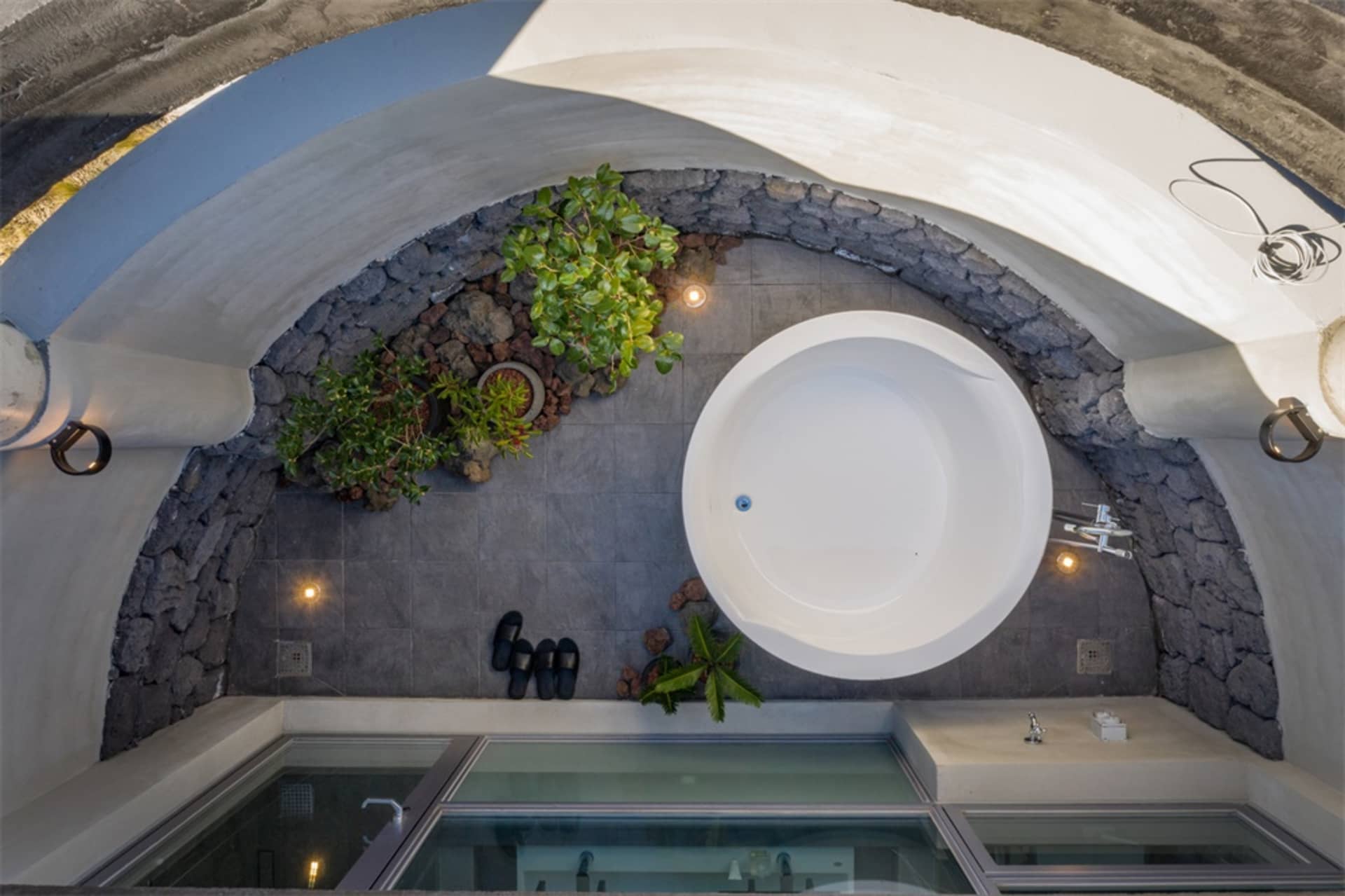 A modern bathroom featuring a circular bathtub surrounded by concrete tiles, plants, and black flip-flops.
