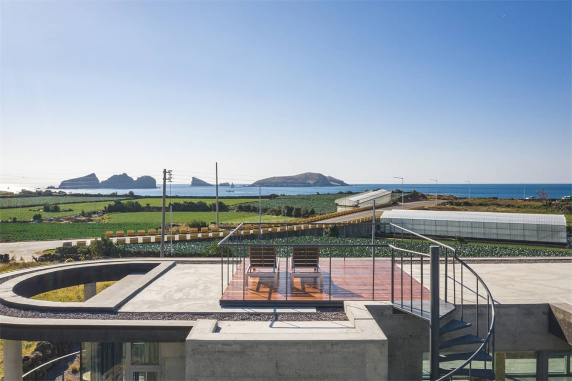 A modern residential exterior featuring a concrete patio with lounge chairs overlooking a lush green field and the ocean.