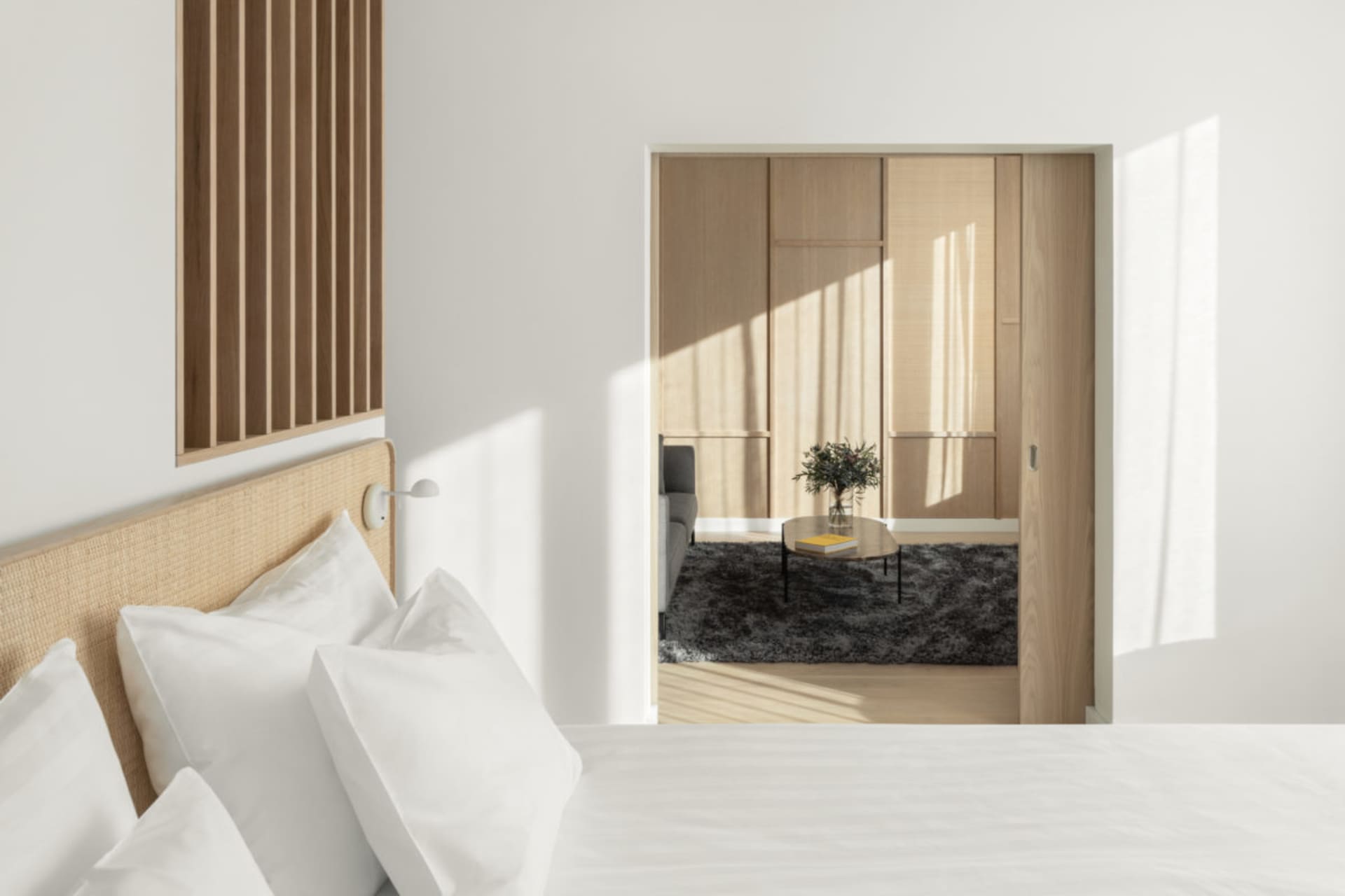 Modern, minimalist bedroom with white bedding, wooden furniture, and light streaming through the window.