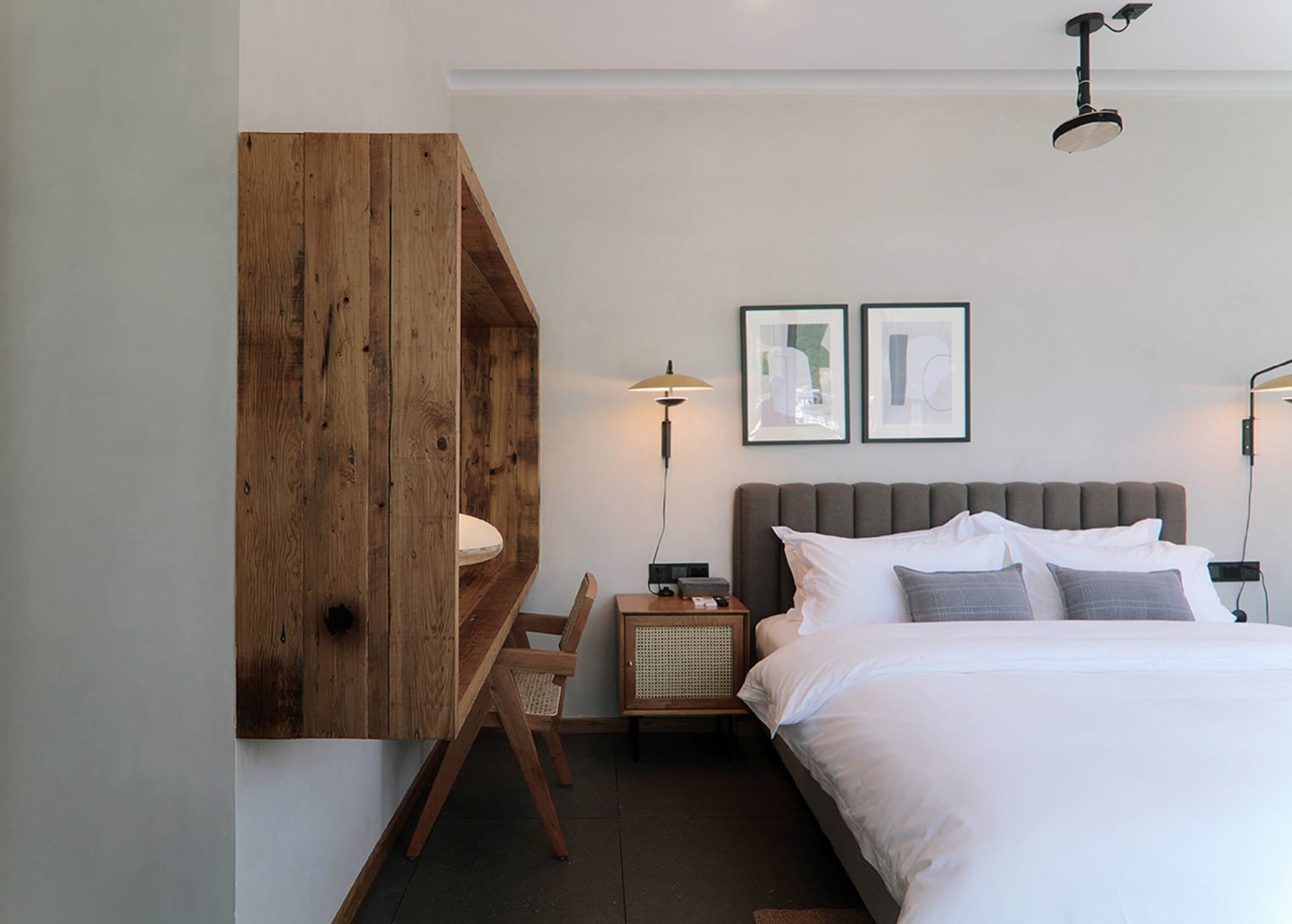 A minimalist bedroom featuring a wooden desk, concrete wall, and white textiles on a large bed.
