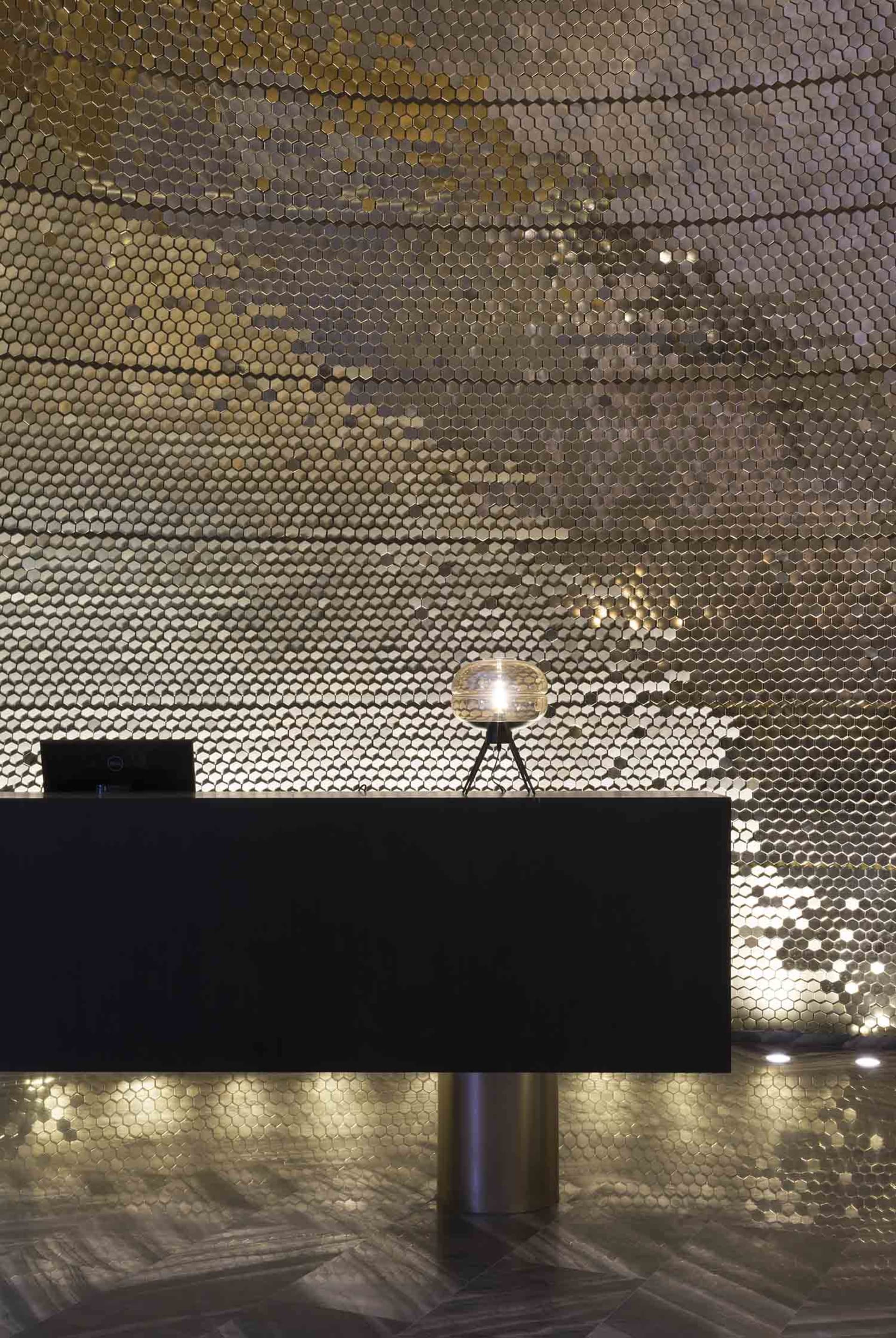 A modern lobby with metallic tiles on the walls and floor, featuring a sleek reception desk with a glass lamp.
