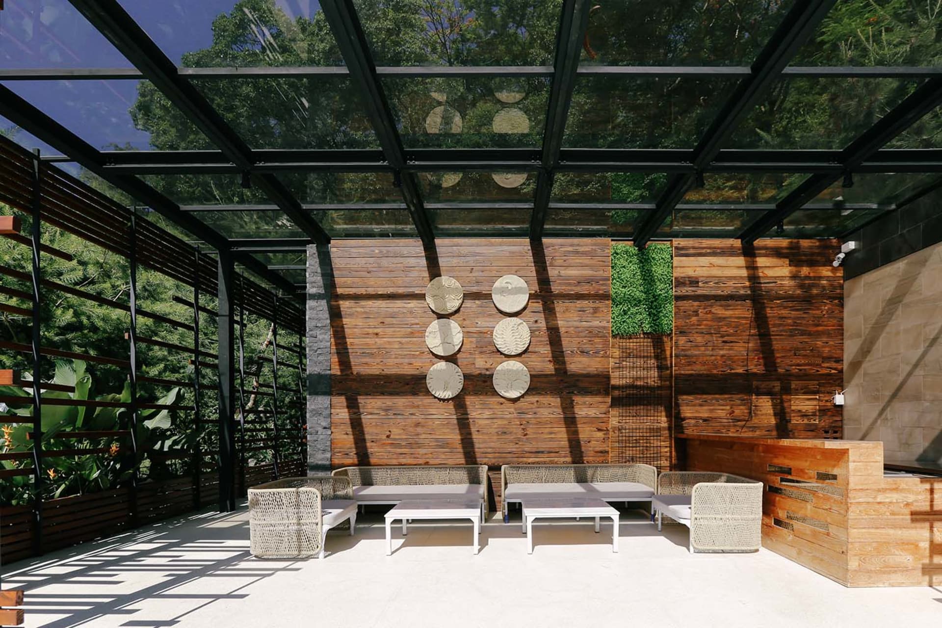 A modern patio lounge area with wooden elements, including a wooden roof supported by glass beams and wooden walls adorned wi