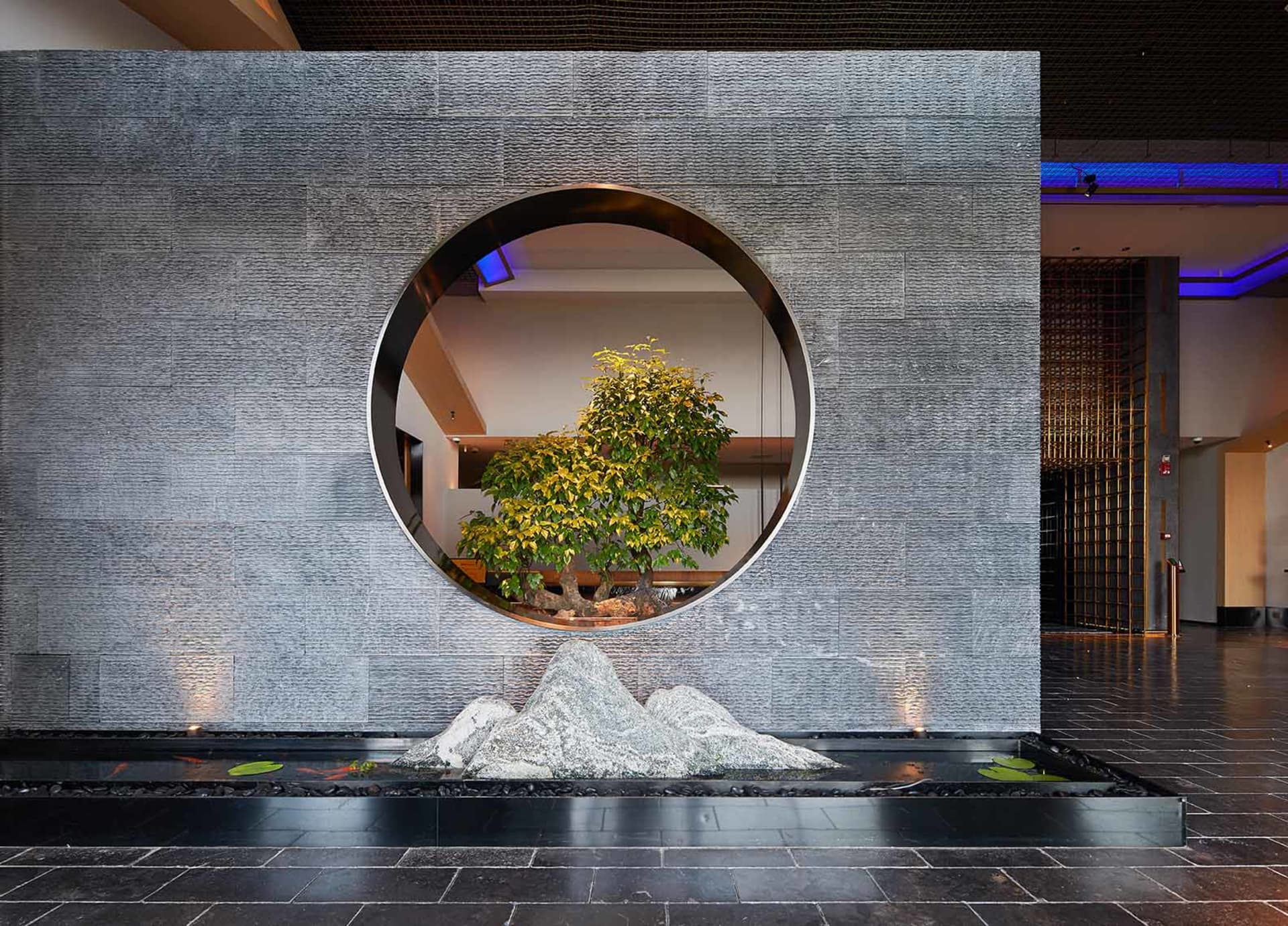 Modern hotel lobby with a circular window, tree, and water feature.