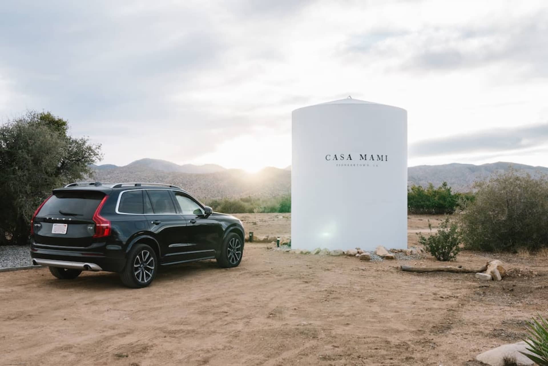 Black SUV parked next to a white cylindrical monument with the words "Casa Mami" on it, against a backdrop of mountains and d