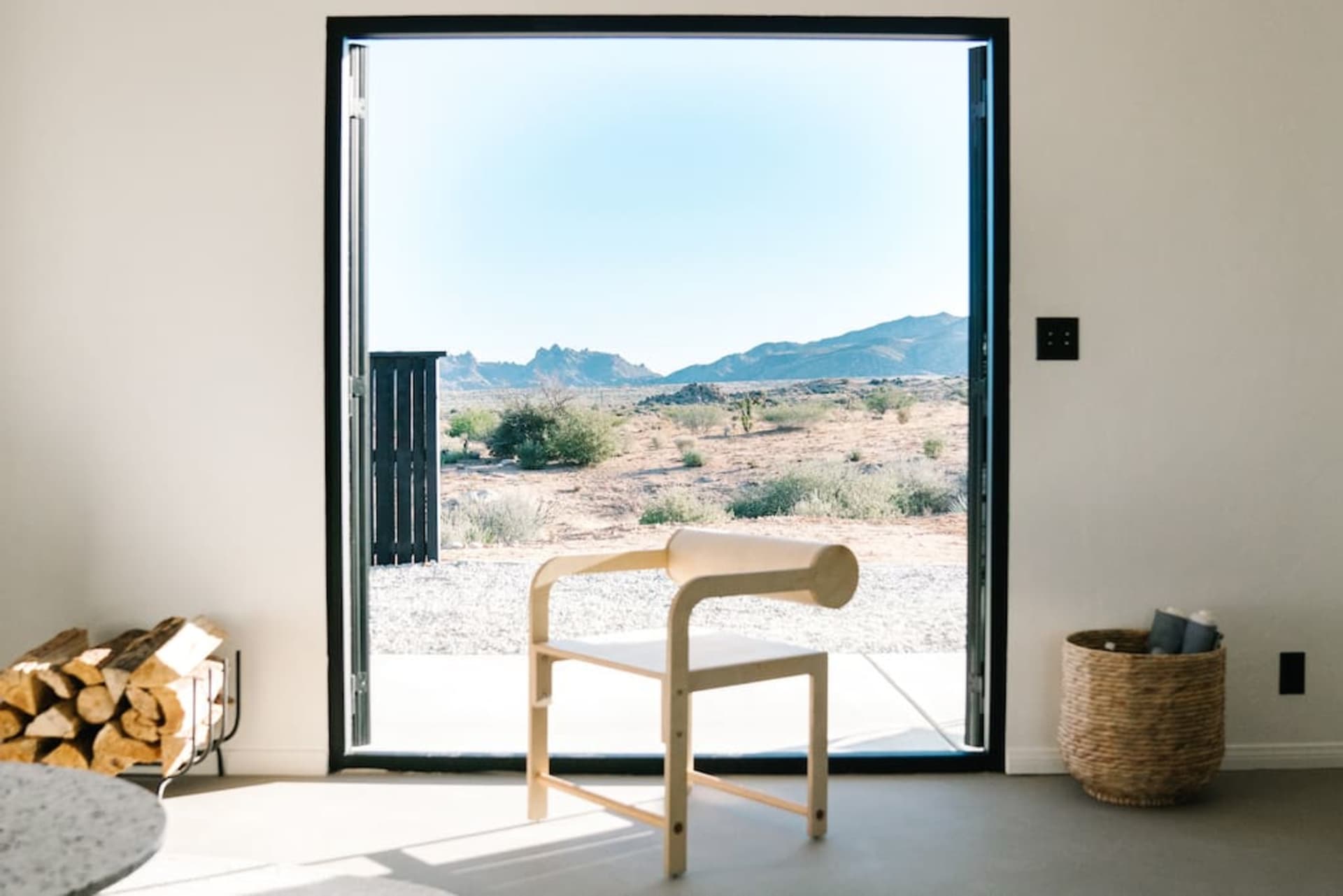 A minimalist living room with a desert view through a large window, featuring a wooden chair, a wicker basket, and a stack of