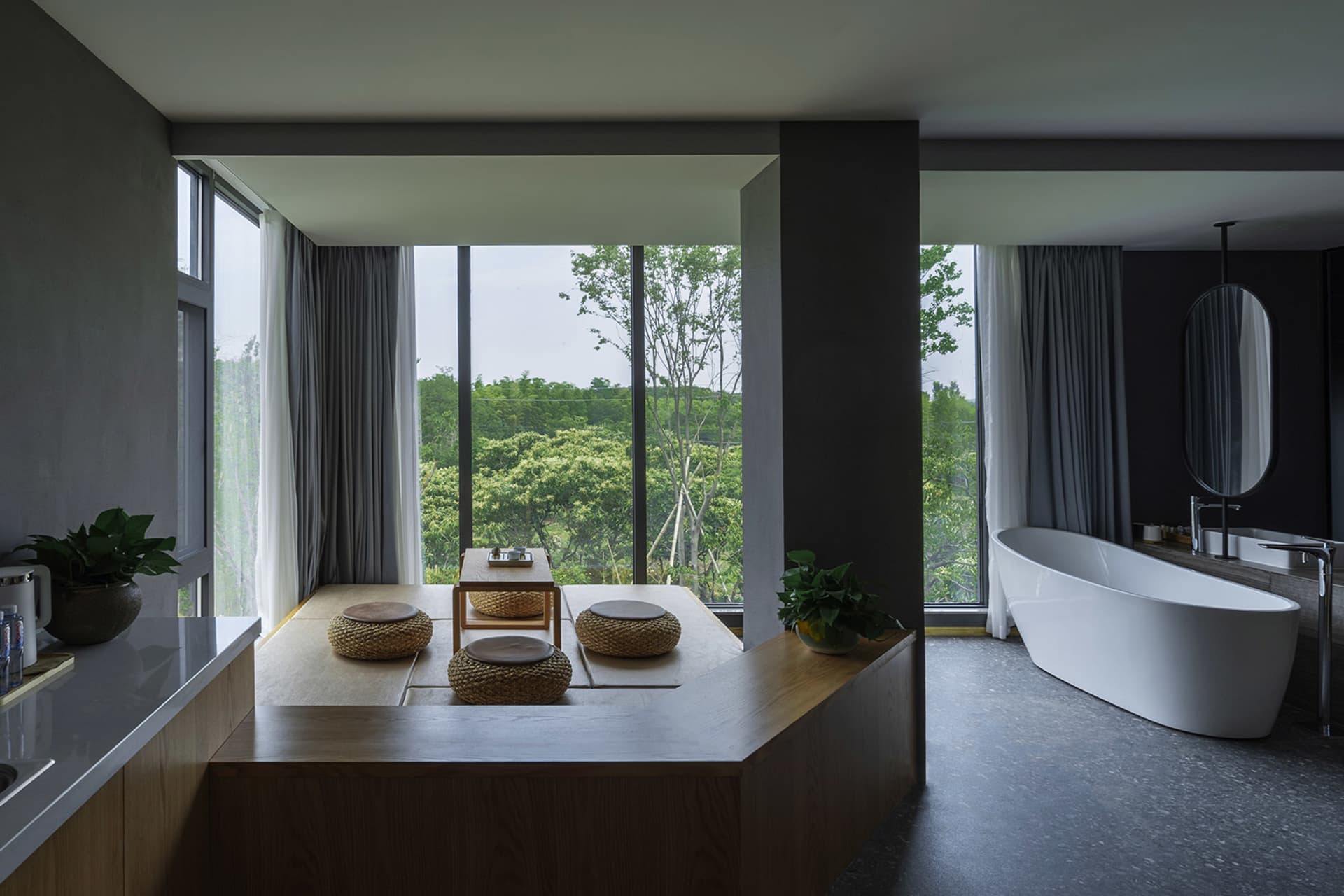 A modern residential bathroom with wood, concrete, and glass elements, featuring a bathtub, seating area, and large windows.