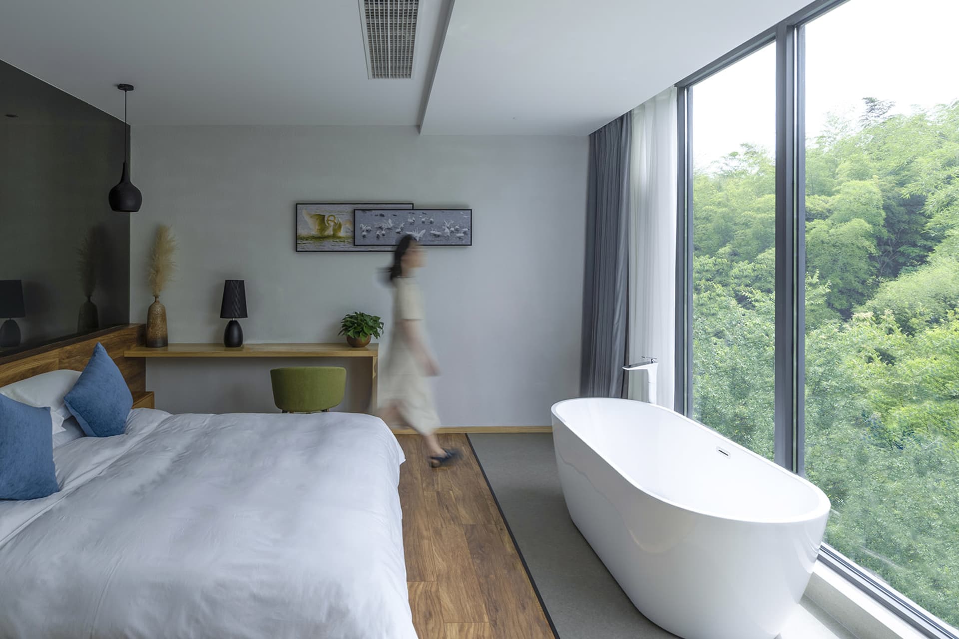 A modern bedroom with a white freestanding bathtub and a large window overlooking greenery.