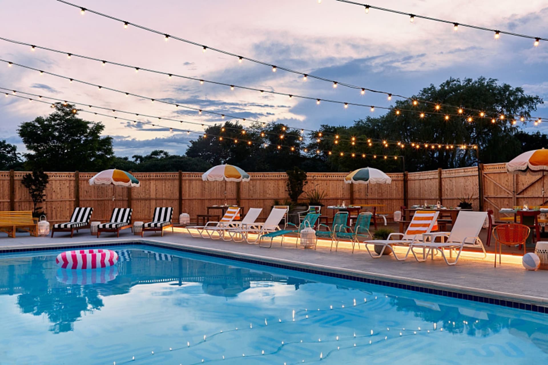 A serene residential backyard with a wooden fence, a sparkling swimming pool, colorful lounge chairs, and string lights.