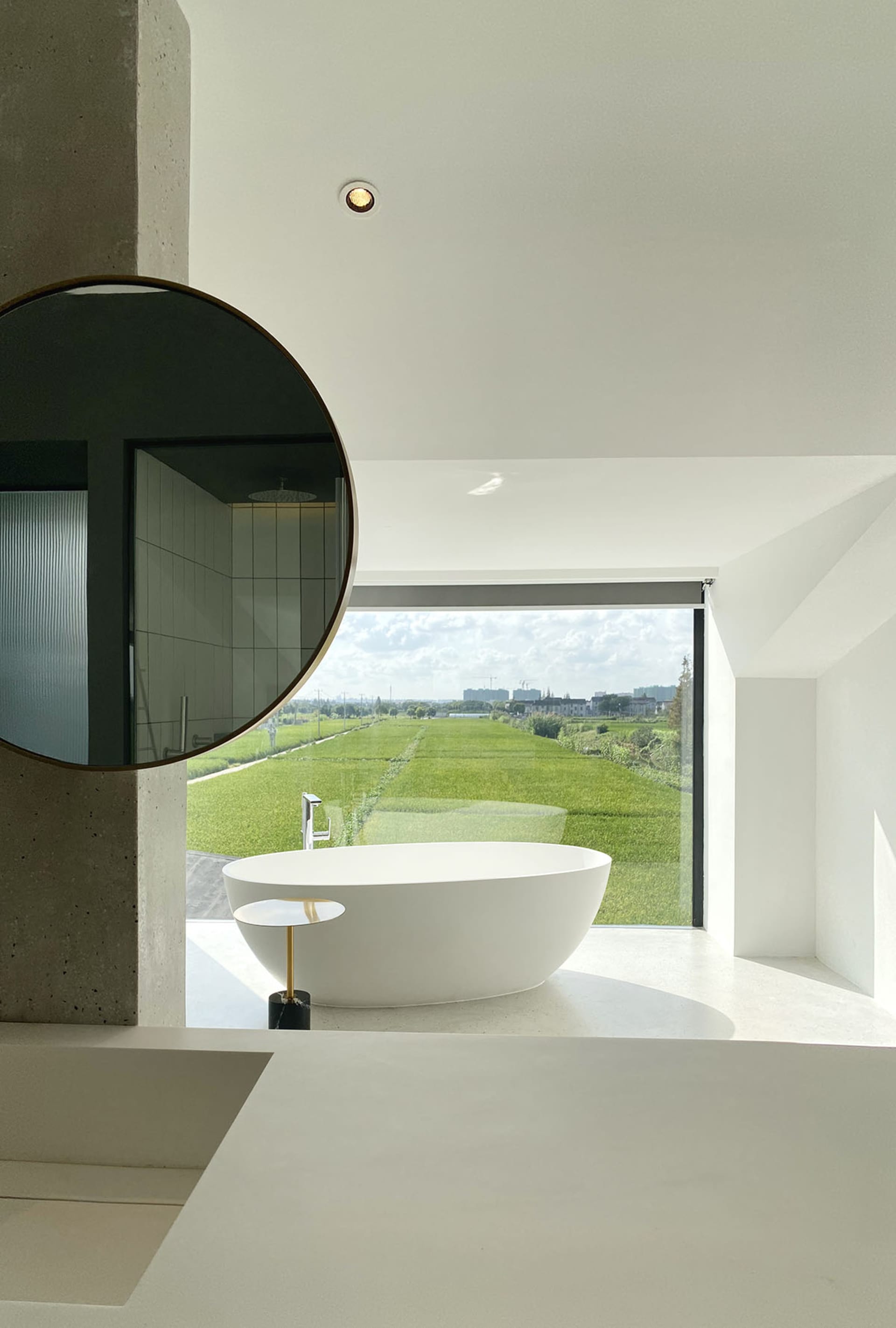 Modern bathroom with a minimalist white concrete bathtub, reflecting the lush greenery outside, and a large circular mirror.