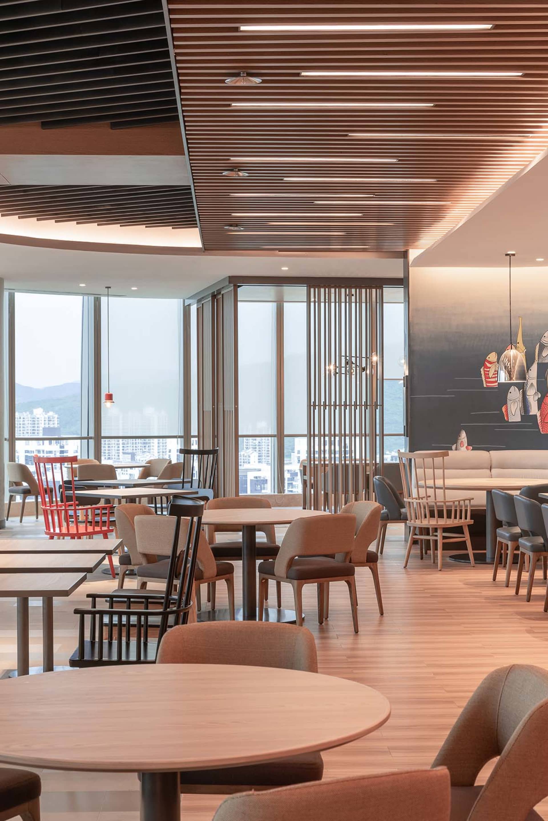 A modern, minimalist restaurant interior with wooden tables and chairs, large windows offering a view of the city, and a warm