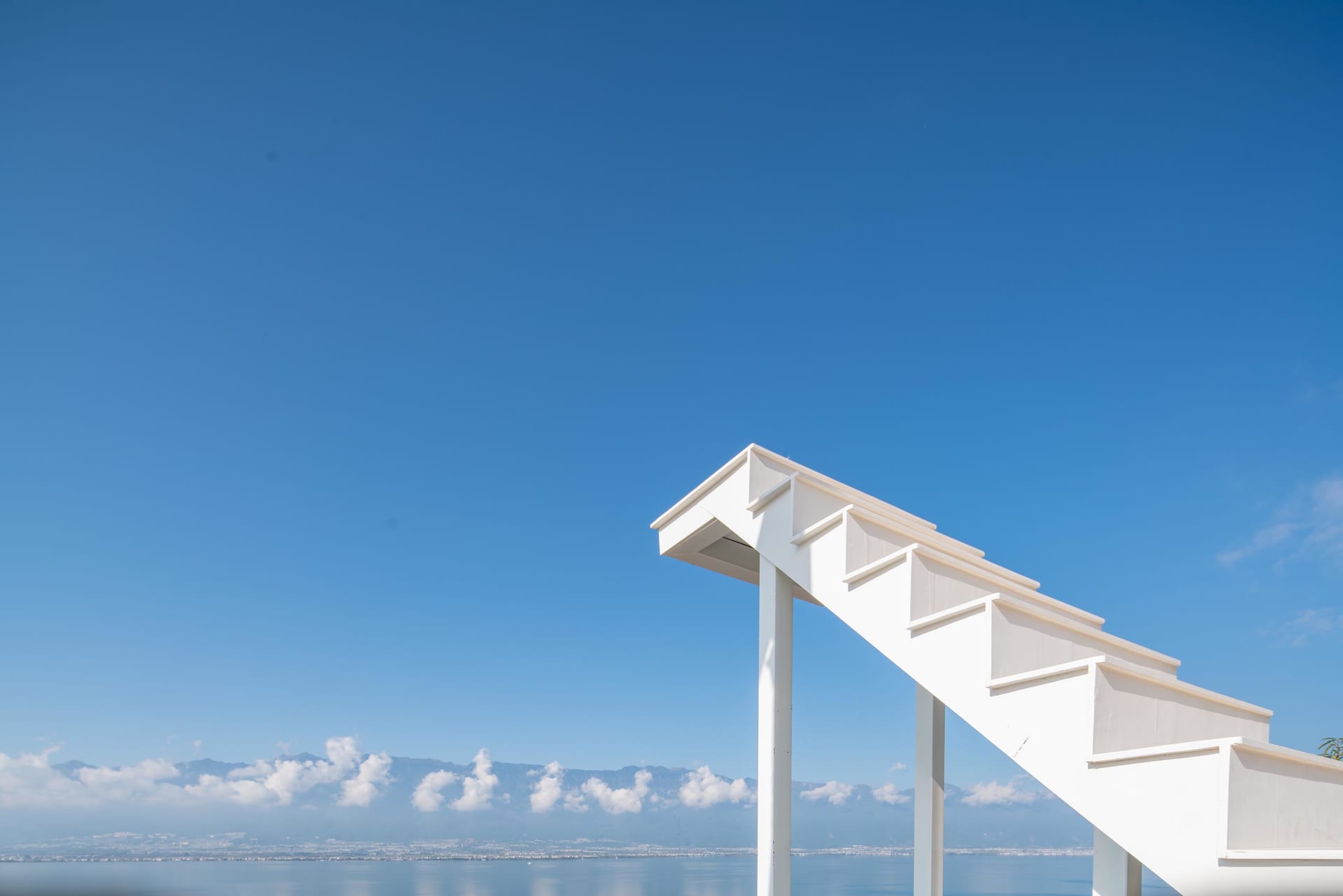 A modern staircase with white painted wooden steps and railings leading down to a beach or waterfront.