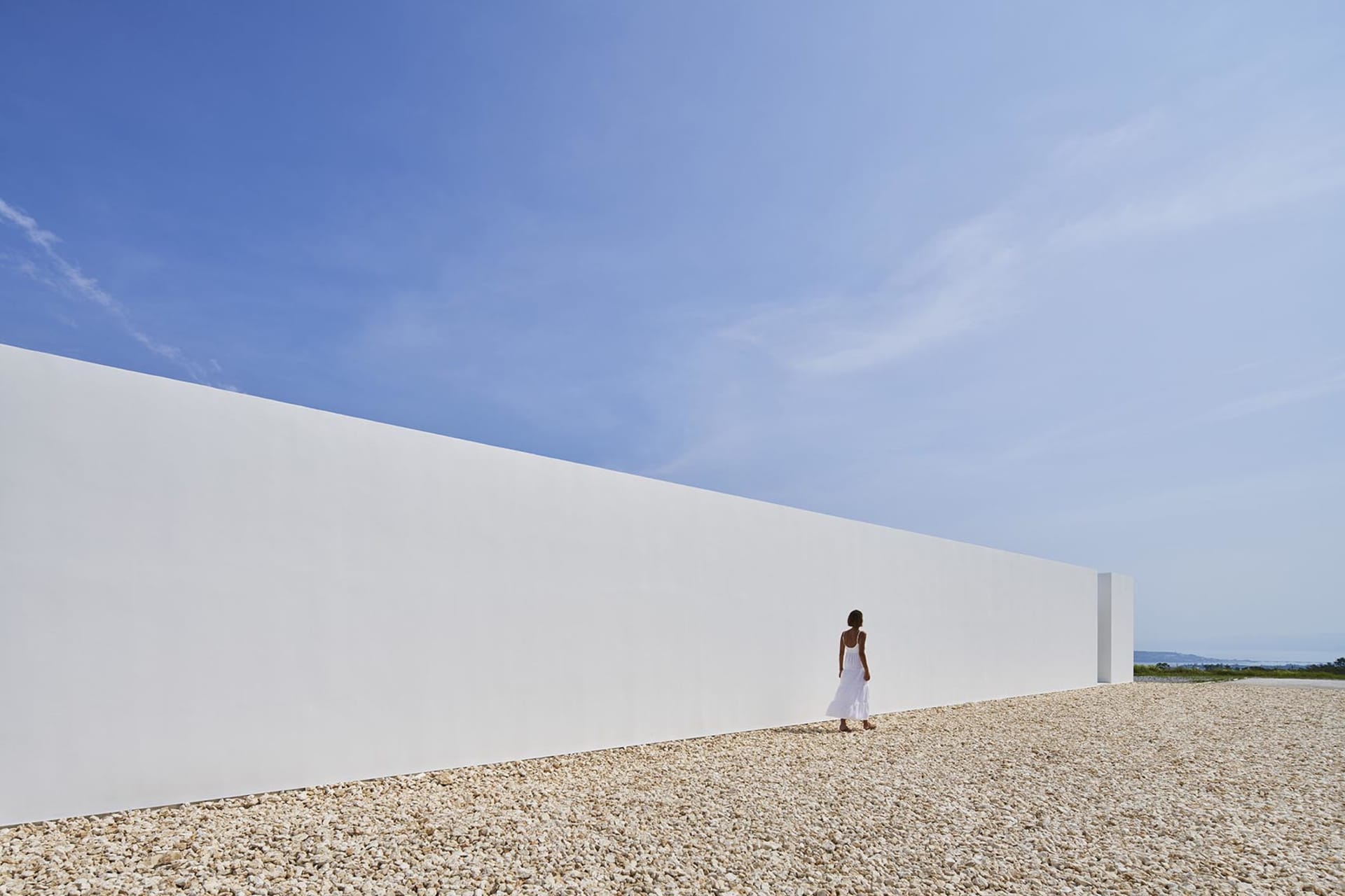 A minimalist white wall along a gravel ground with clear blue sky in the background.