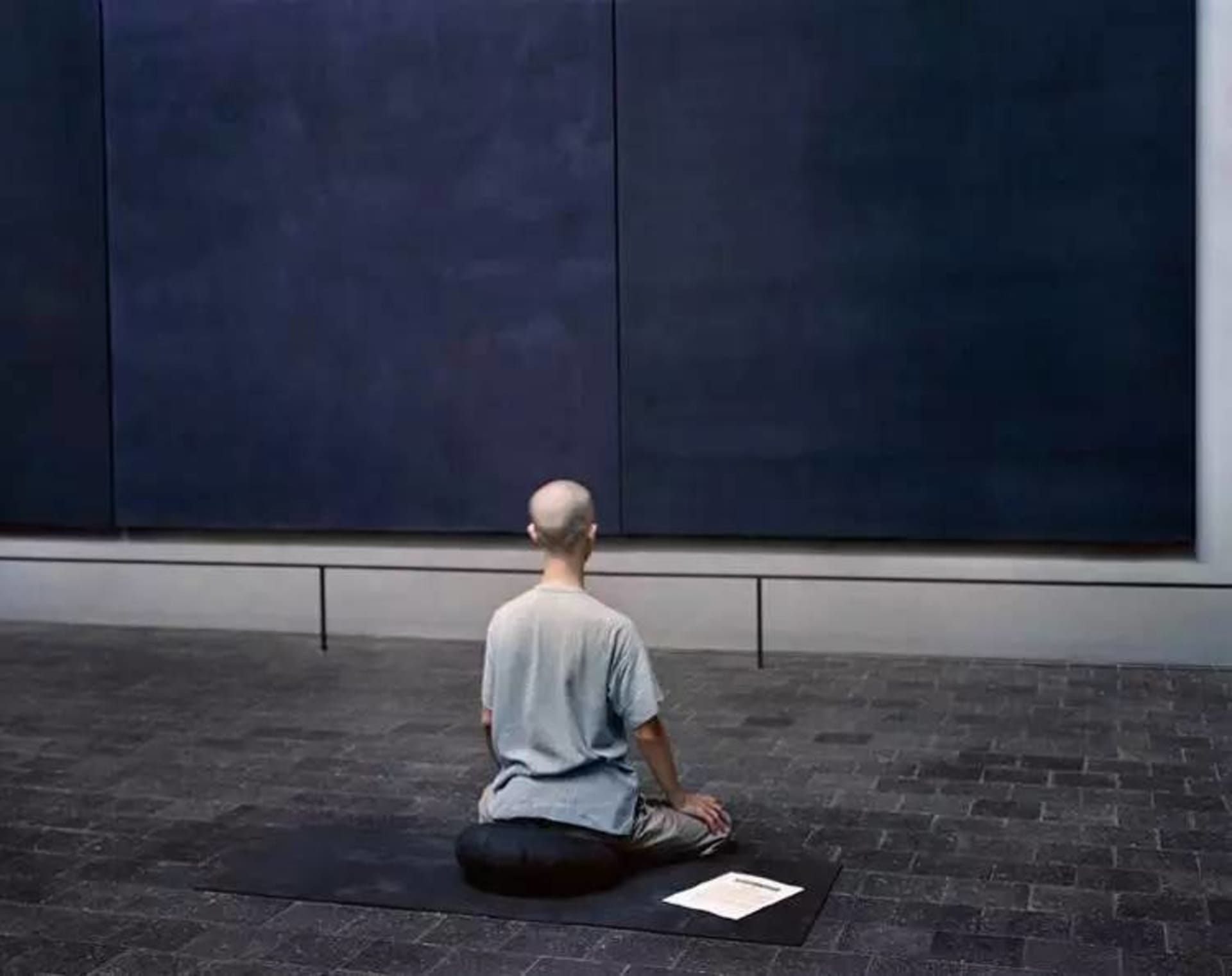 A minimalist installation art piece consisting of a person sitting on a cork mat and looking at a piece of paper in a museum