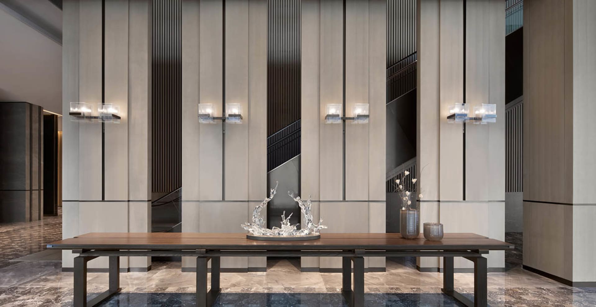 A modern lobby features a long wooden table with metal legs, adorned with a glass water feature, set against a backdrop of ma