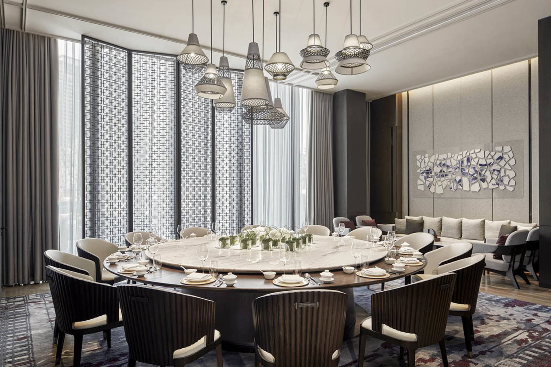 A modern dining room featuring a large, round marble table surrounded by high-backed chairs, with pendant lights hanging abov