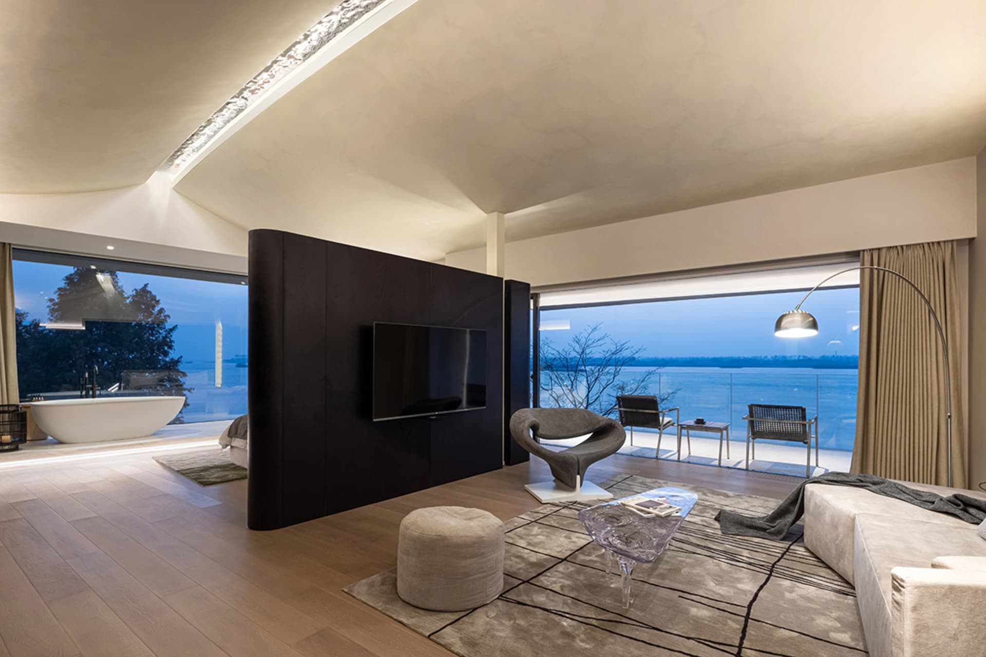 A modern living room with large windows overlooking a body of water, featuring minimalist furniture and a television mounted