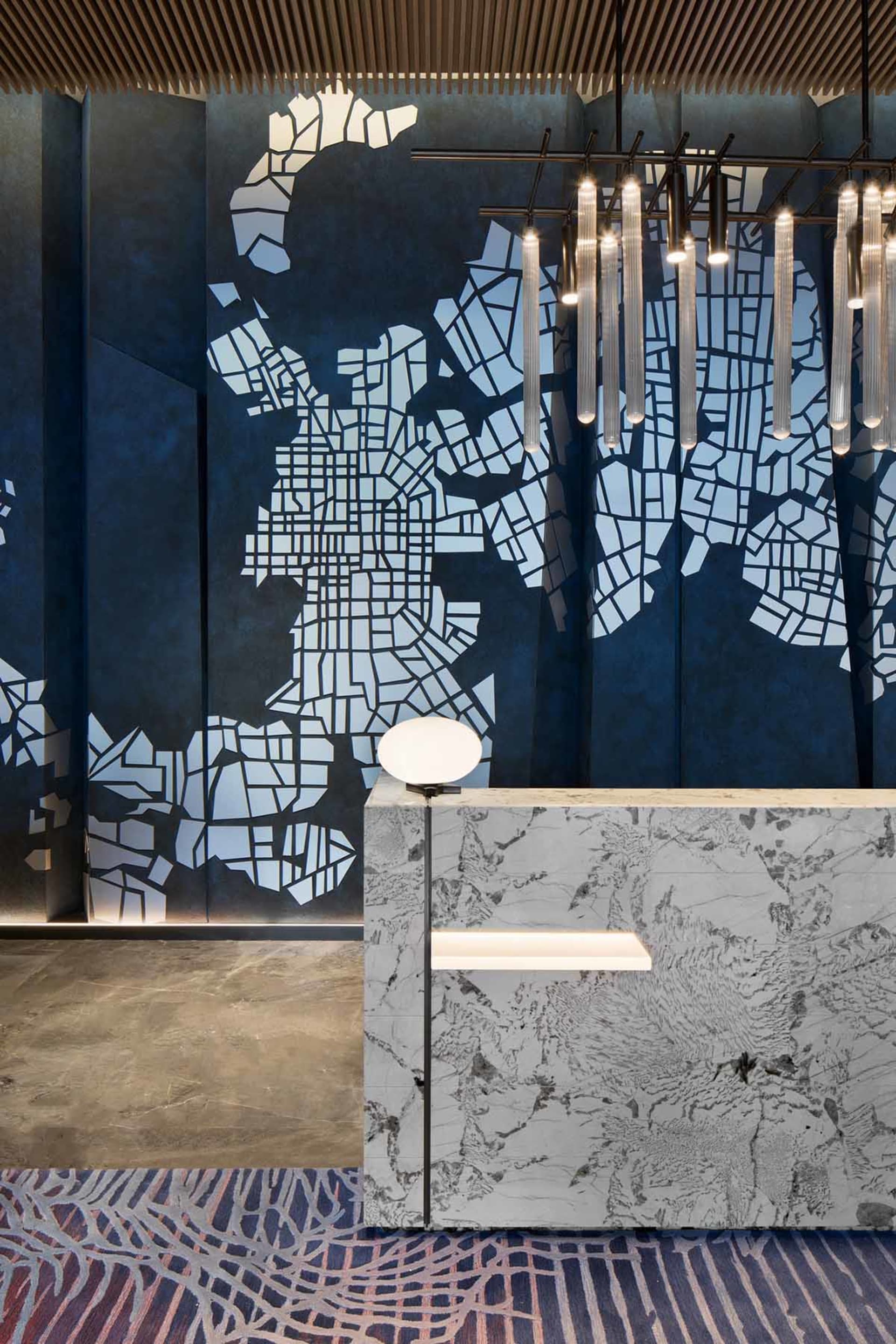 A modern lobby featuring a marble wall with a map pattern, ambient lighting, and geometric hanging lights.