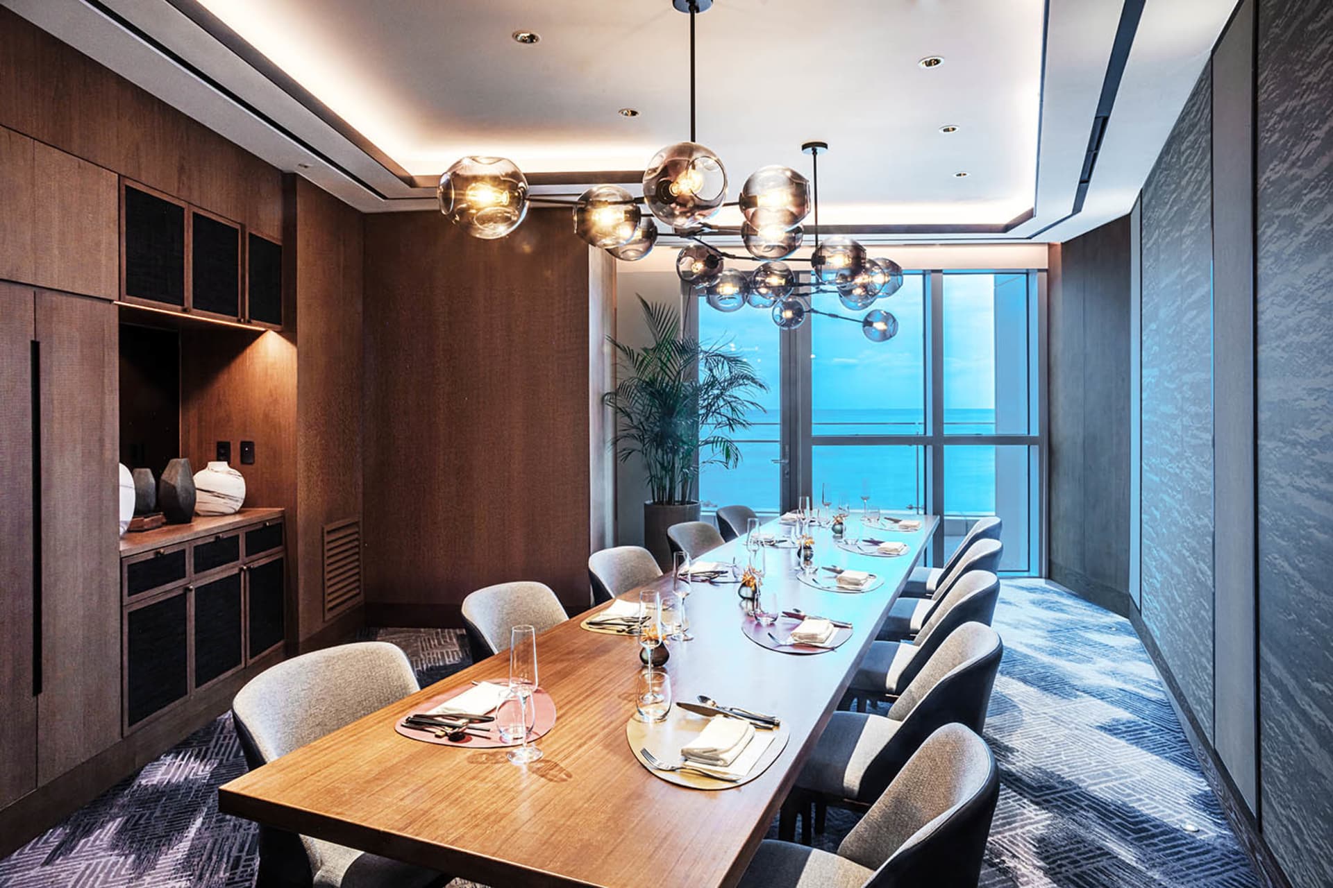 A modern restaurant dining room featuring a wooden table and chairs, chandelier, and ocean view.