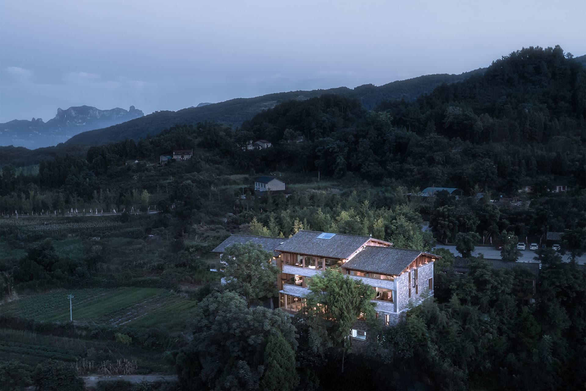 A modern residential building with a wooden roof and windows, surrounded by greenery and mountains.