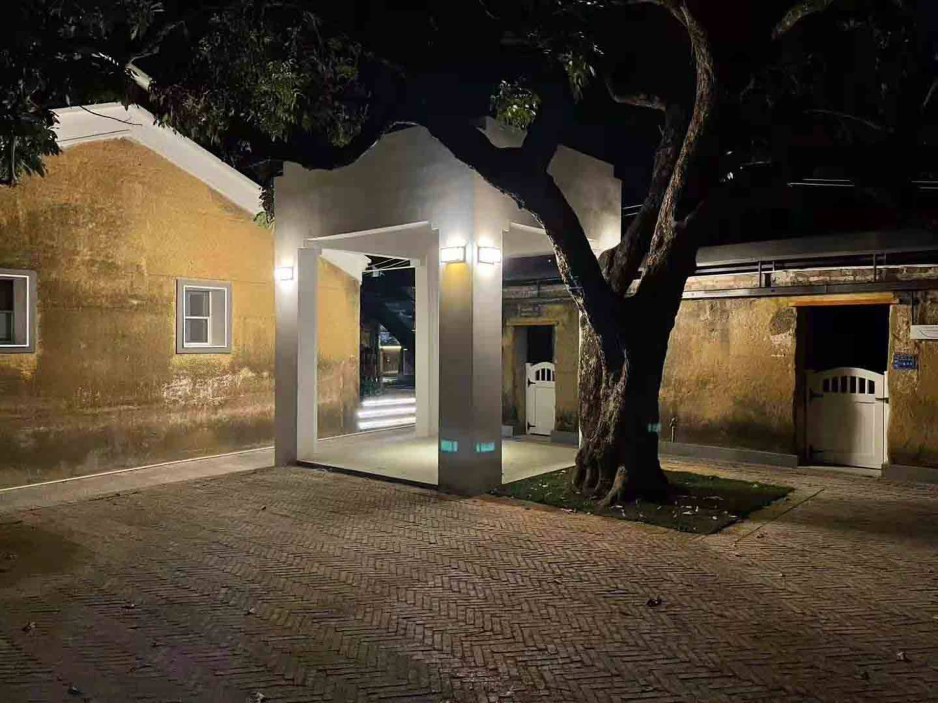 極簡玄關以木質立面搭配白牆，夜晚在大型樹木下由暖光照亮。