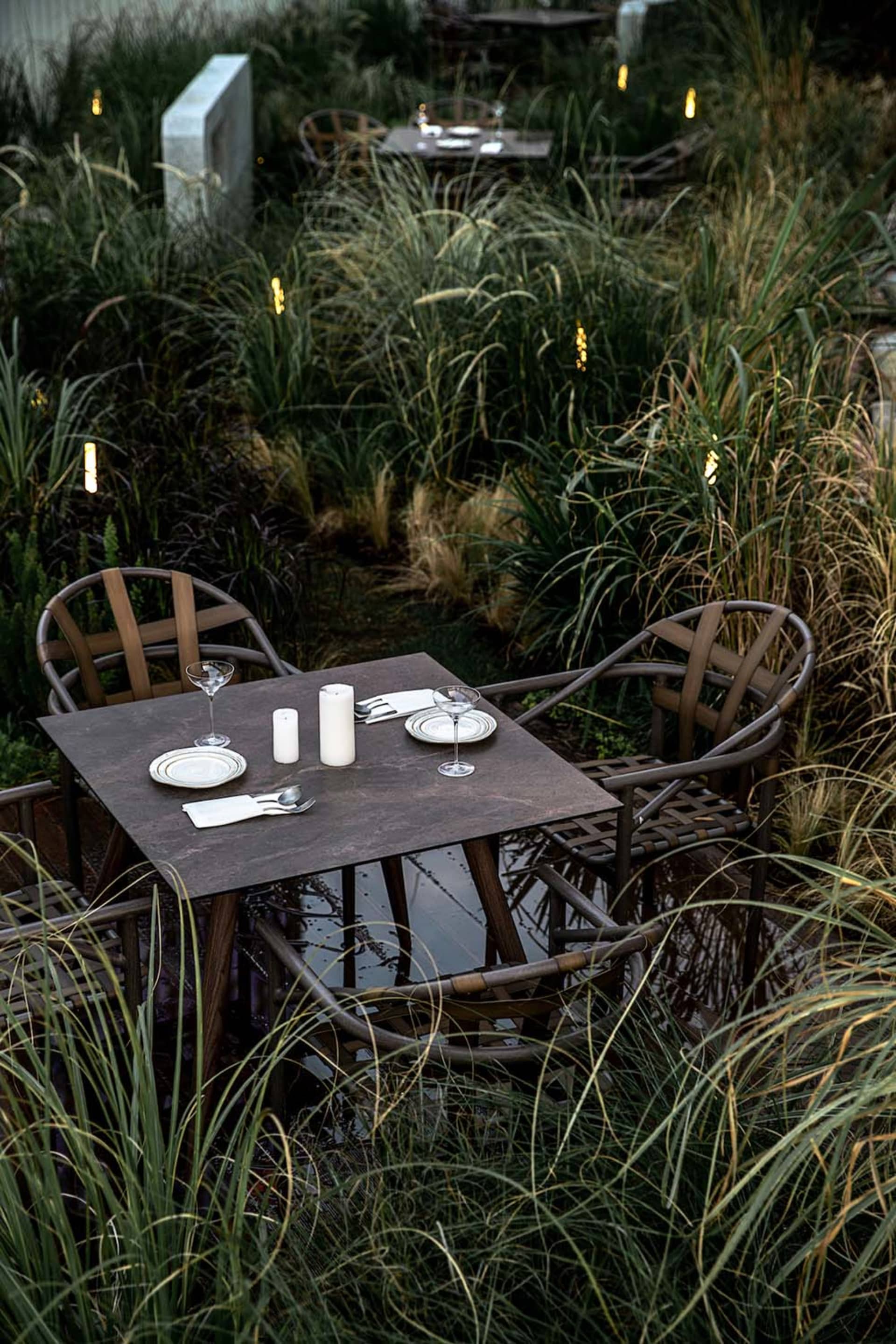 Modern outdoor dining area with metal chairs and glass table surrounded by tall grasses and illuminated by string lights.