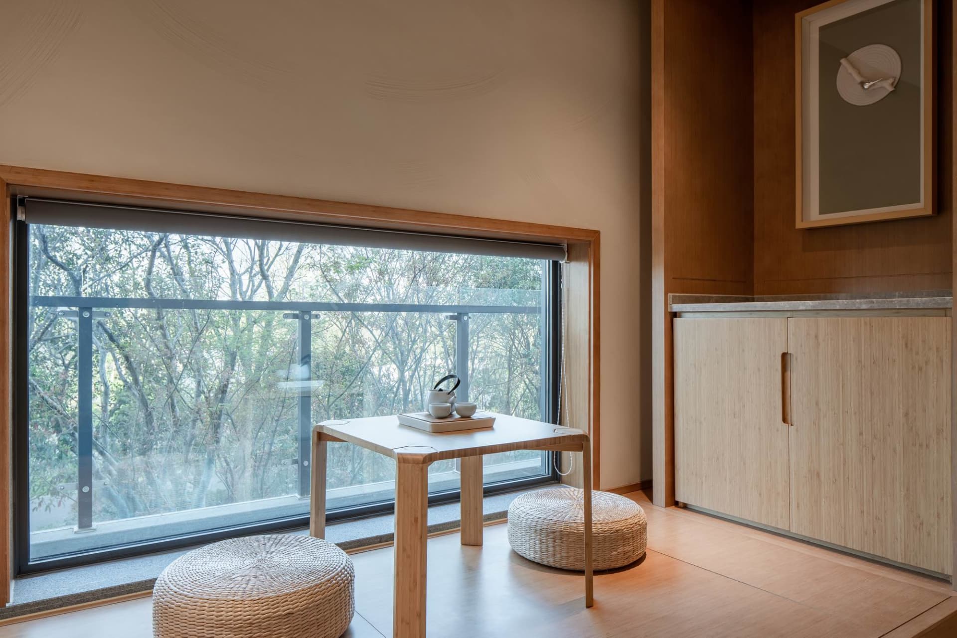 A modern tea room featuring woven stools, a wooden table, and a large window overlooking the trees.