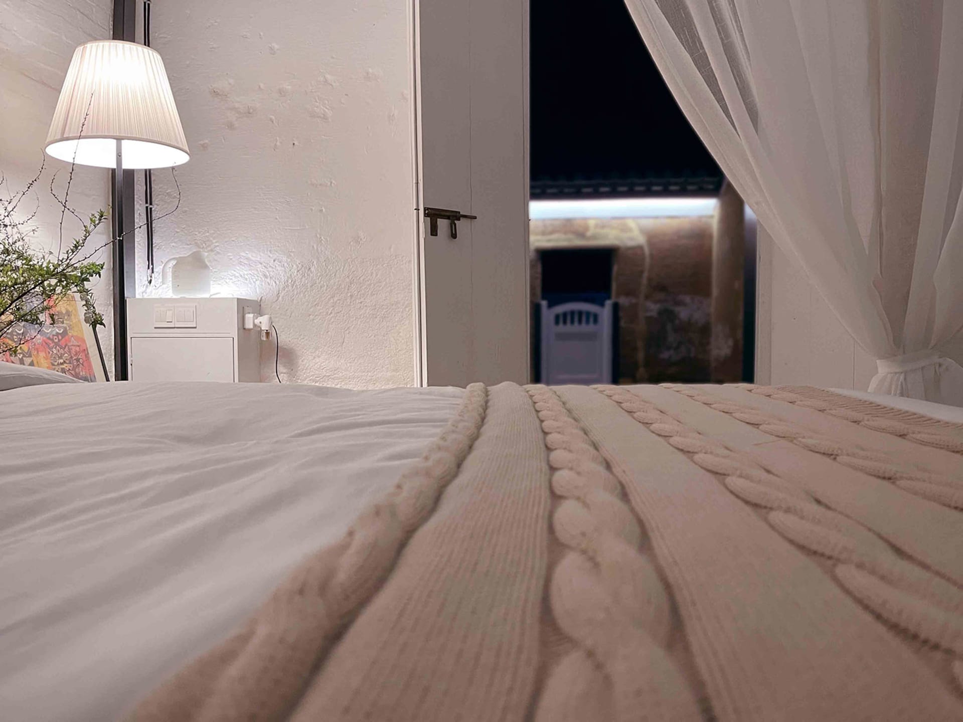 A neatly made bed with a textured white bedspread, lamp illuminating the room, and a view through the open curtains to an out