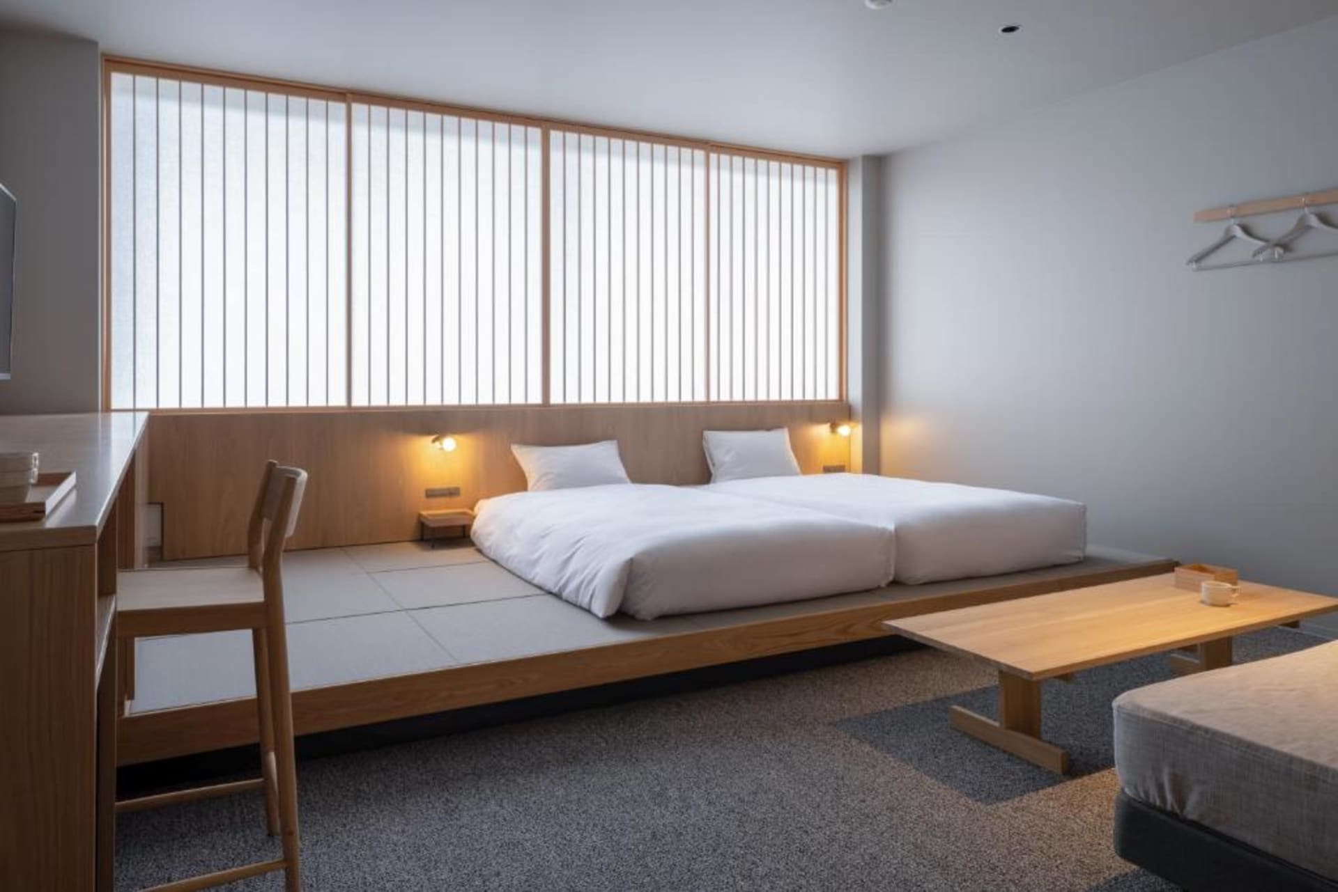 A minimalist bedroom featuring white bedding on a low wooden platform bed, a simple wooden table, and bright natural light en