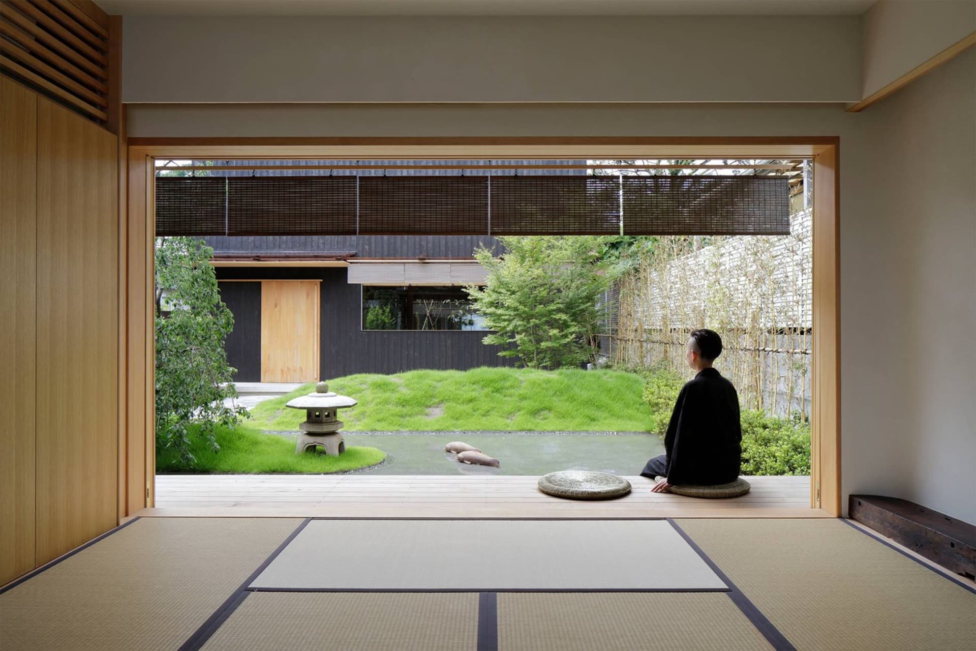 Person in traditional Japanese attire sitting on a tatami mat, looking out at a traditional Japanese garden with a stone lant