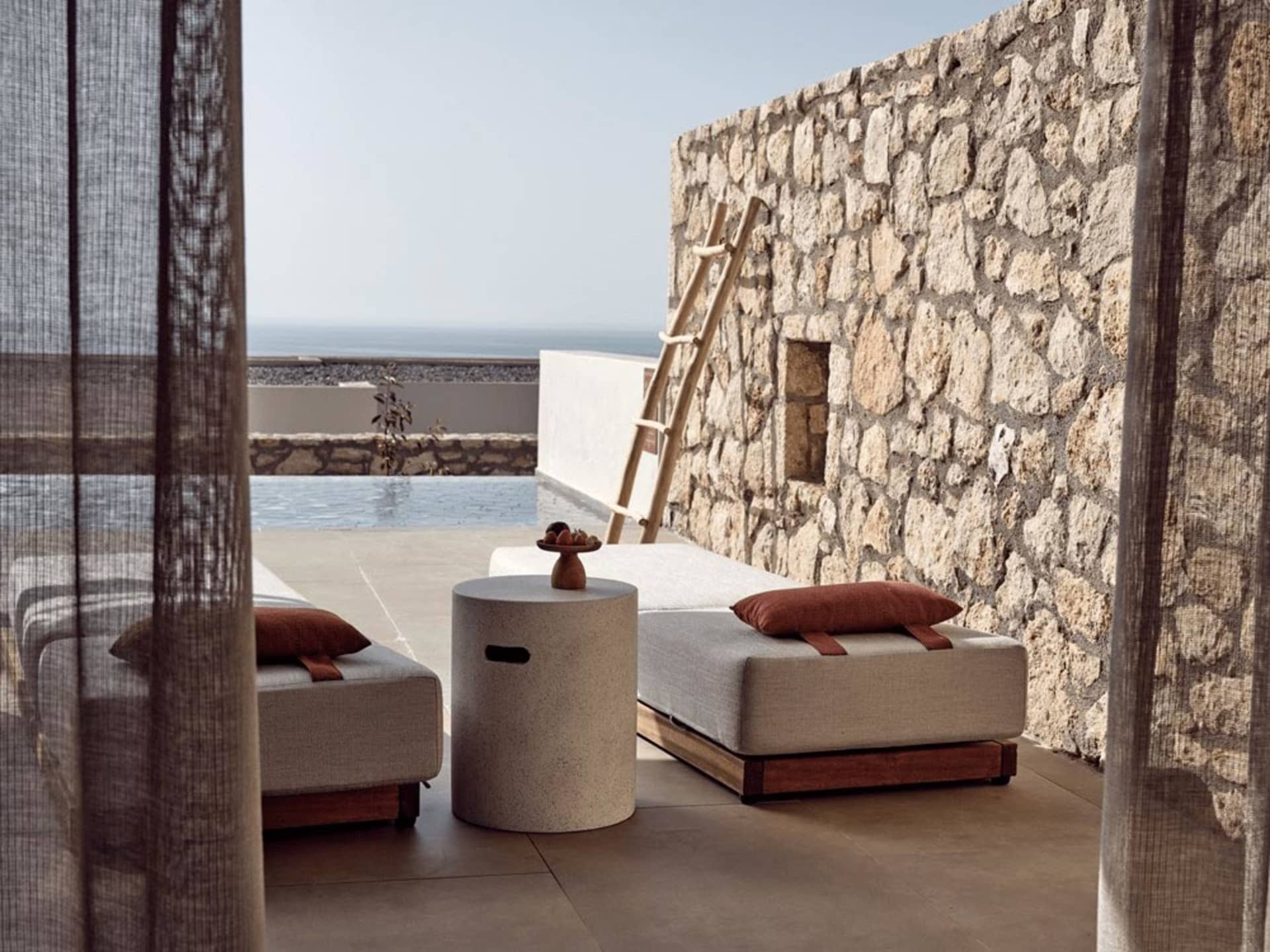 Modern outdoor lounge furniture including plush seating, stone wall, and sea view.