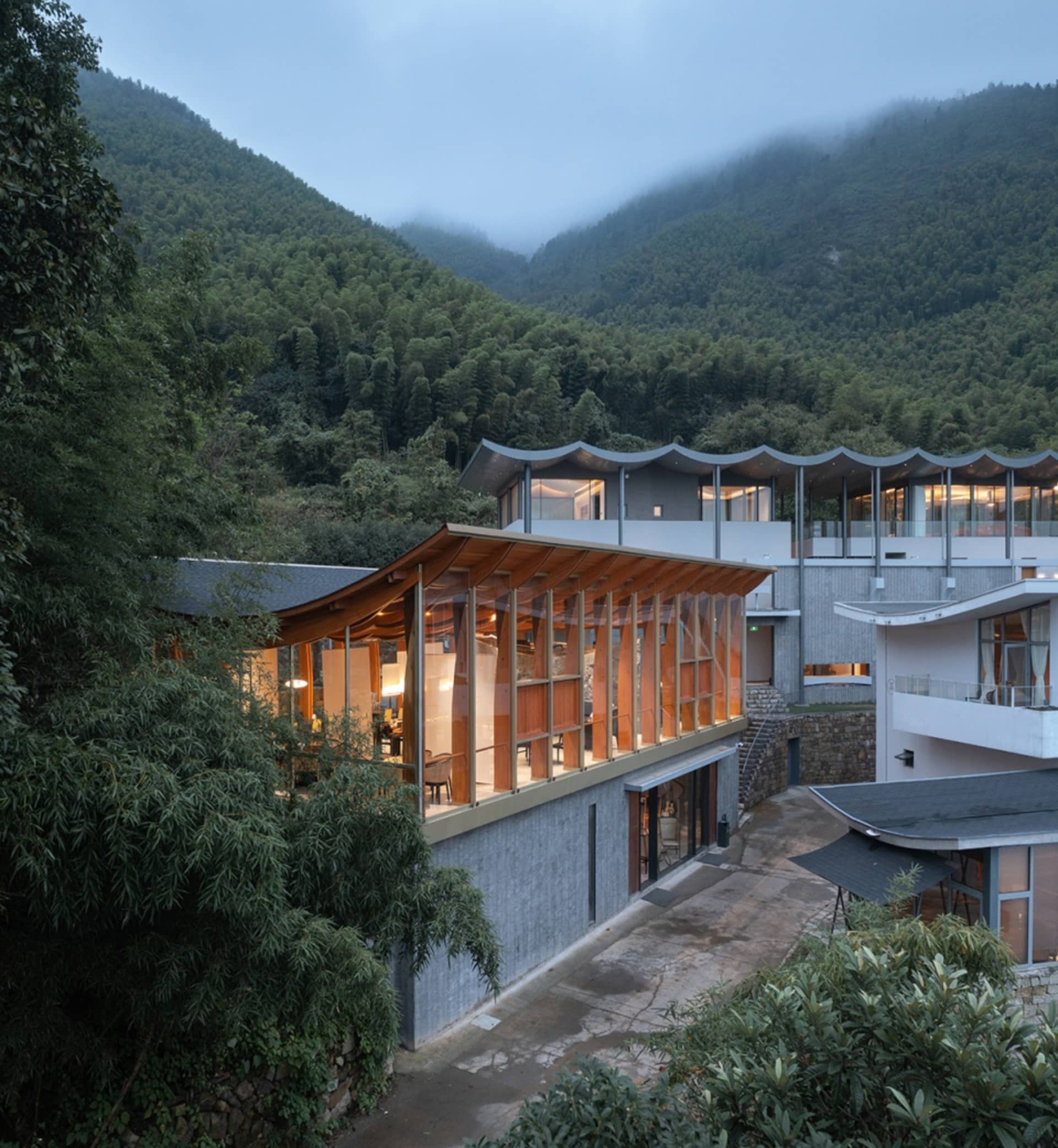 一座以玻璃與混凝土材質為主的現代住宅，周圍環繞茂密綠意，搭配波浪形屋頂與開放式設計