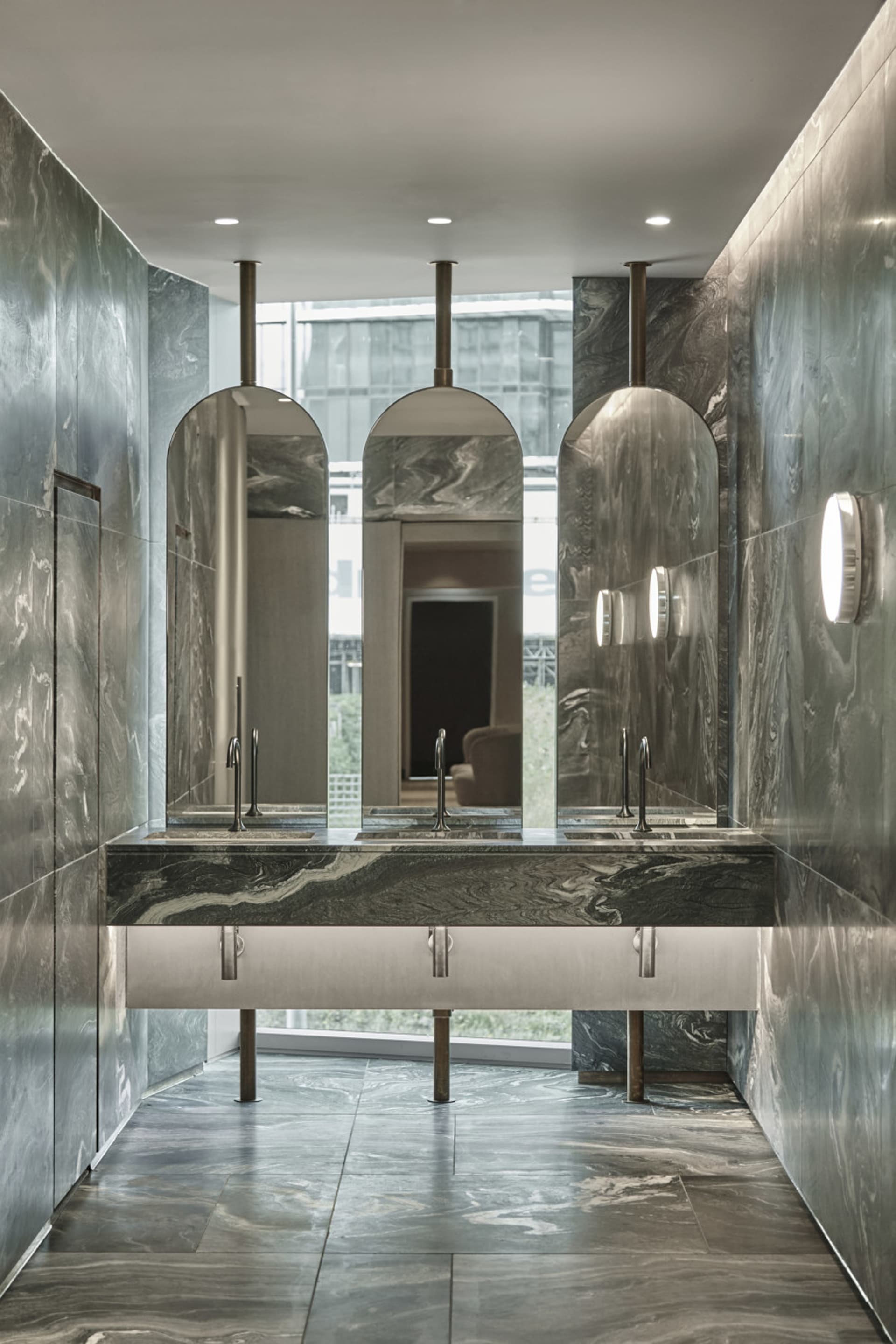 A modern public restroom with marble walls and flooring, featuring a sleek design with arch-shaped mirrors and metal fixtures