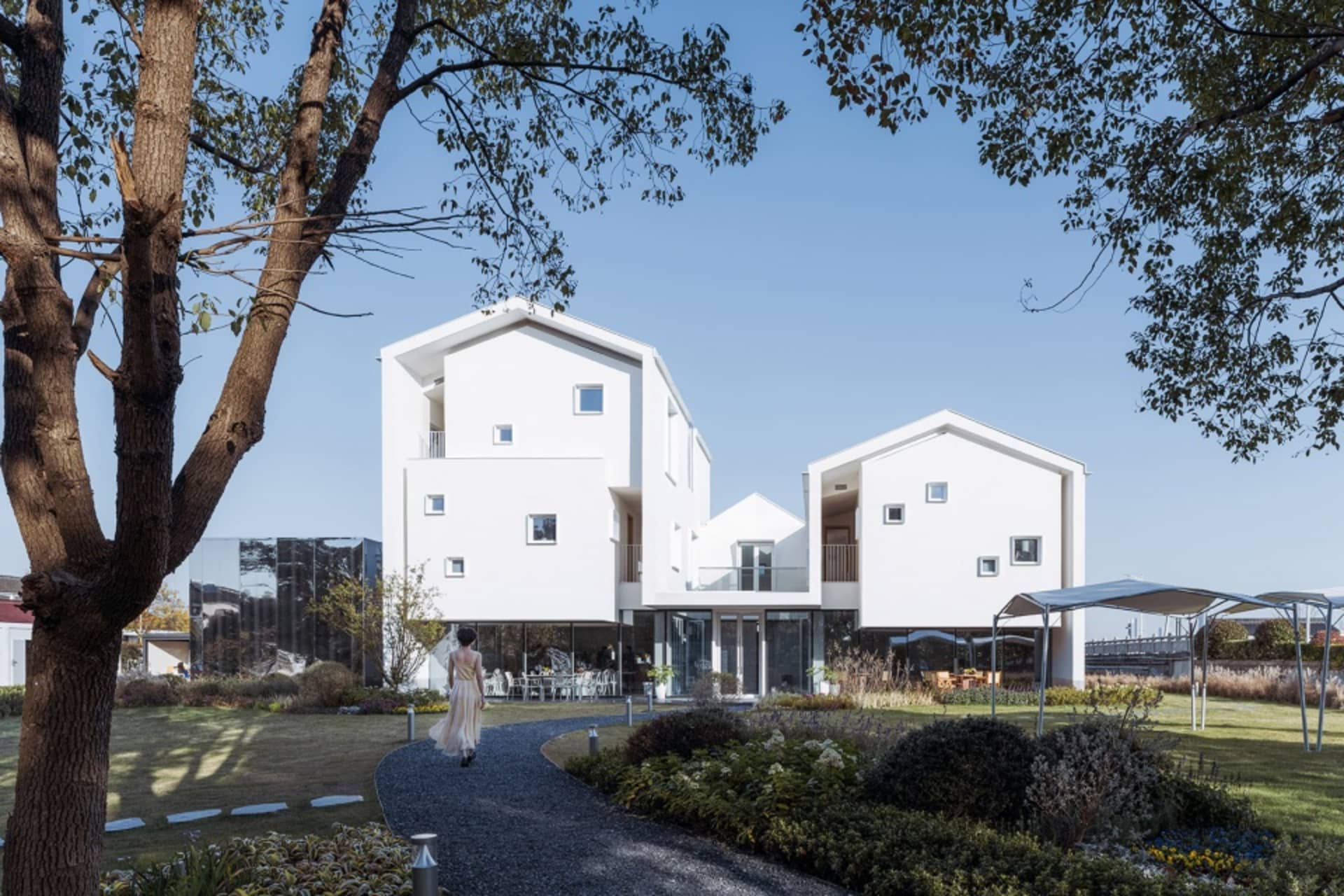 A modern white house with a metal and glass exterior surrounded by greenery and a pathway.