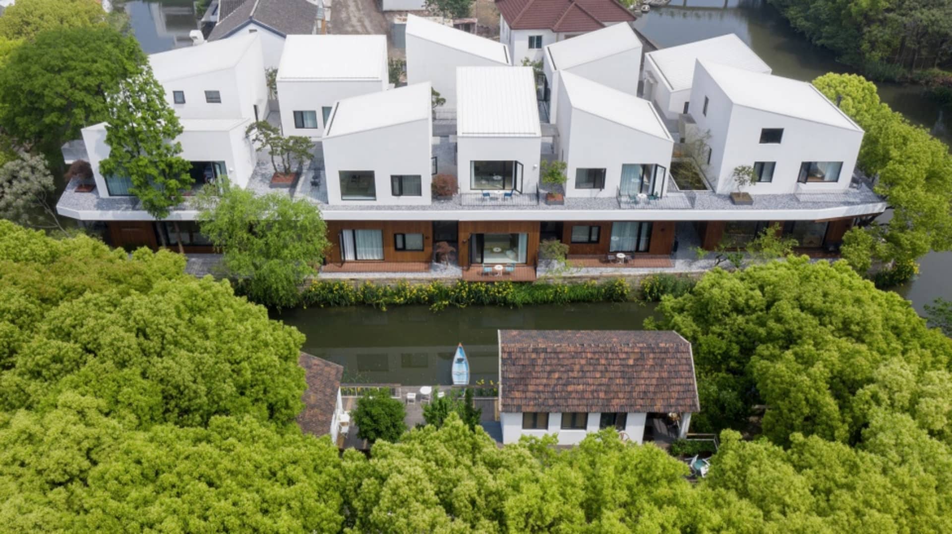 Aerial view of modern black-roof houses with white stucco exteriors and geometric designs, surrounded by greenery and water.