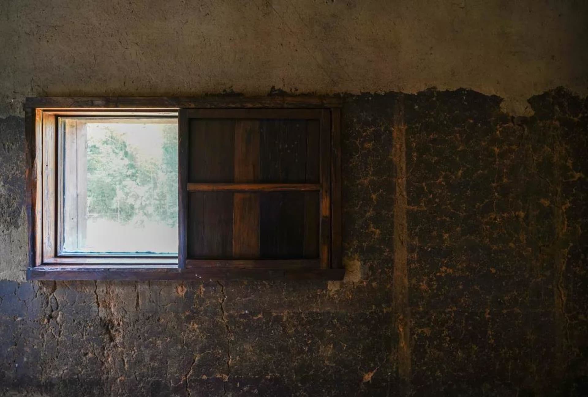 An old, wooden window with a view of greenery outside, set in a cracked, rustic wall.