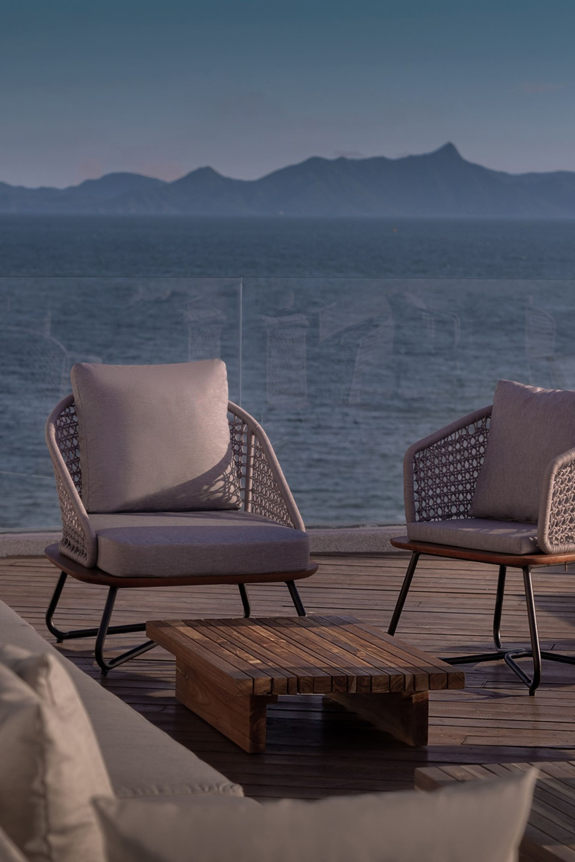 Outdoor seating arrangement featuring wooden furniture and cushions near the ocean with mountains in the background.