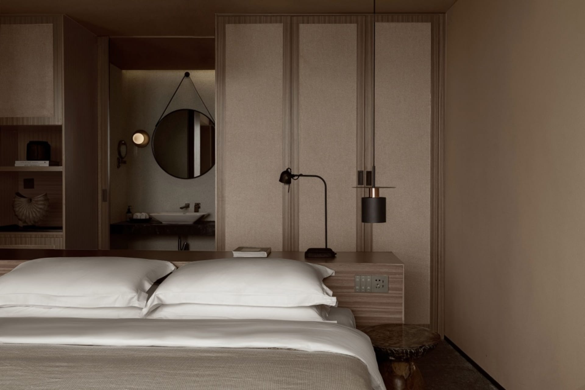 Modern minimalist bedroom with white bedding, wooden furniture, and a round mirror.